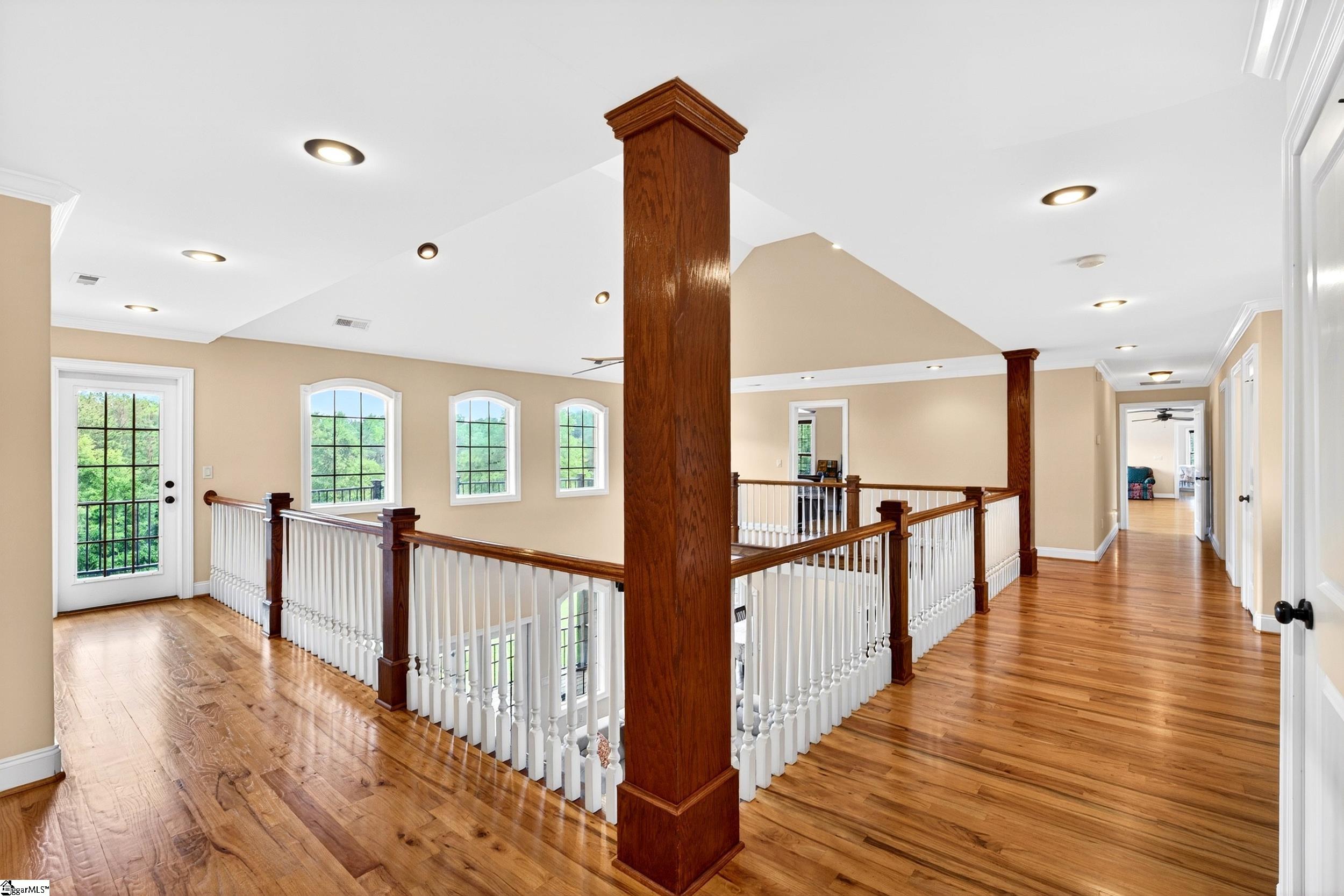 Upstairs landing with hardwood floors, white railings, dark wood columns, and windows overlooking greenery.