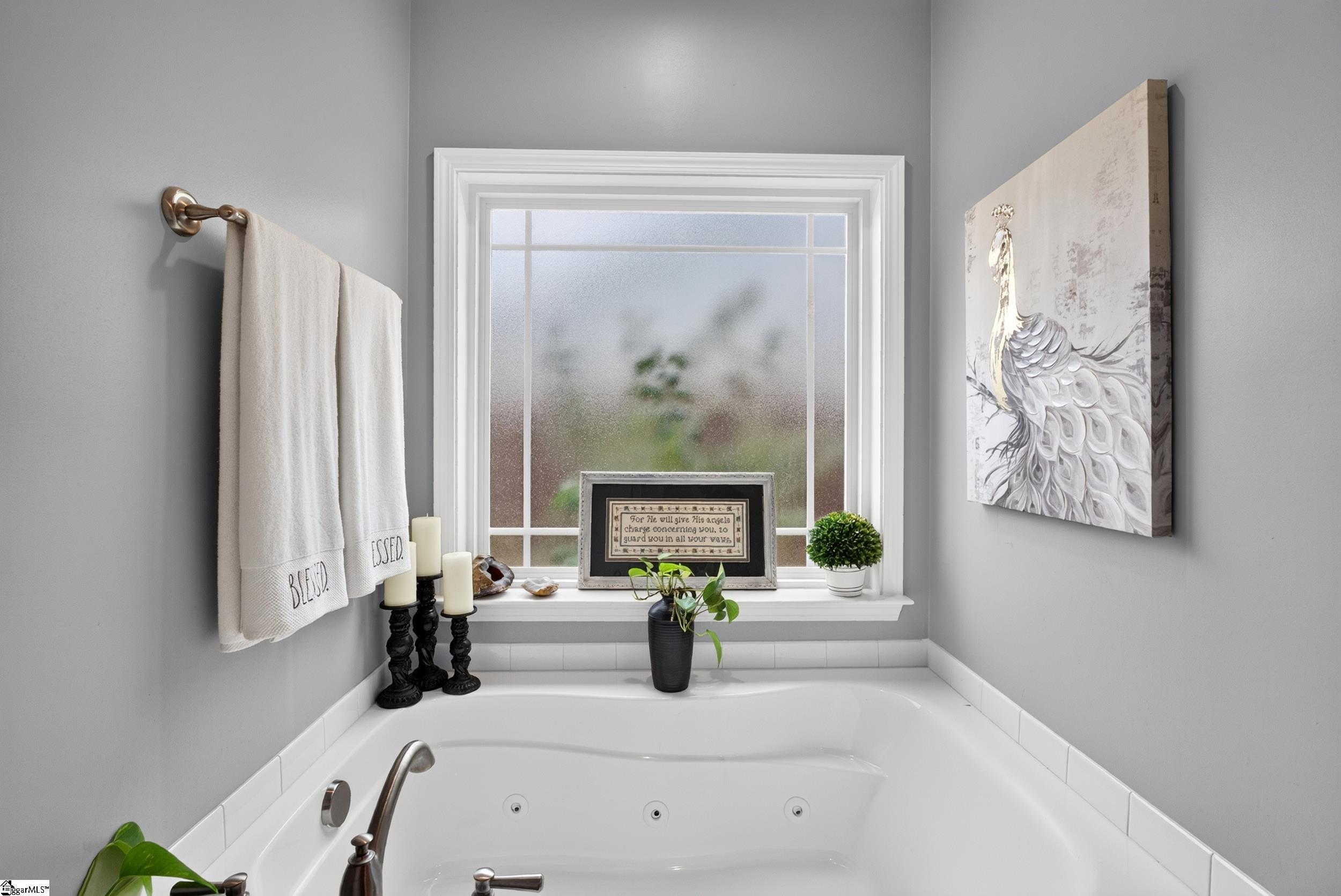 Bathroom jetted tub beneath a large window, decorated with white towels, candles, plants, and framed artwork.