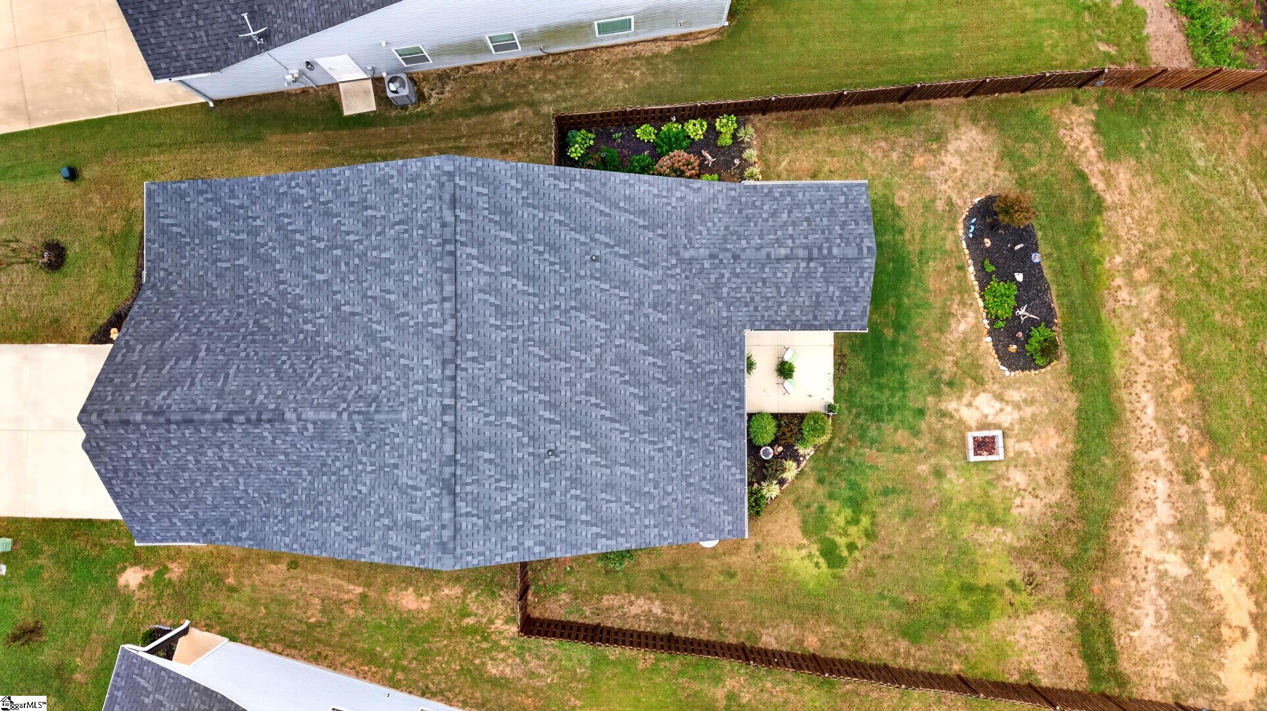 Aerial view of a home with a broad gray roof, fenced yard, garden beds, and a small patio.