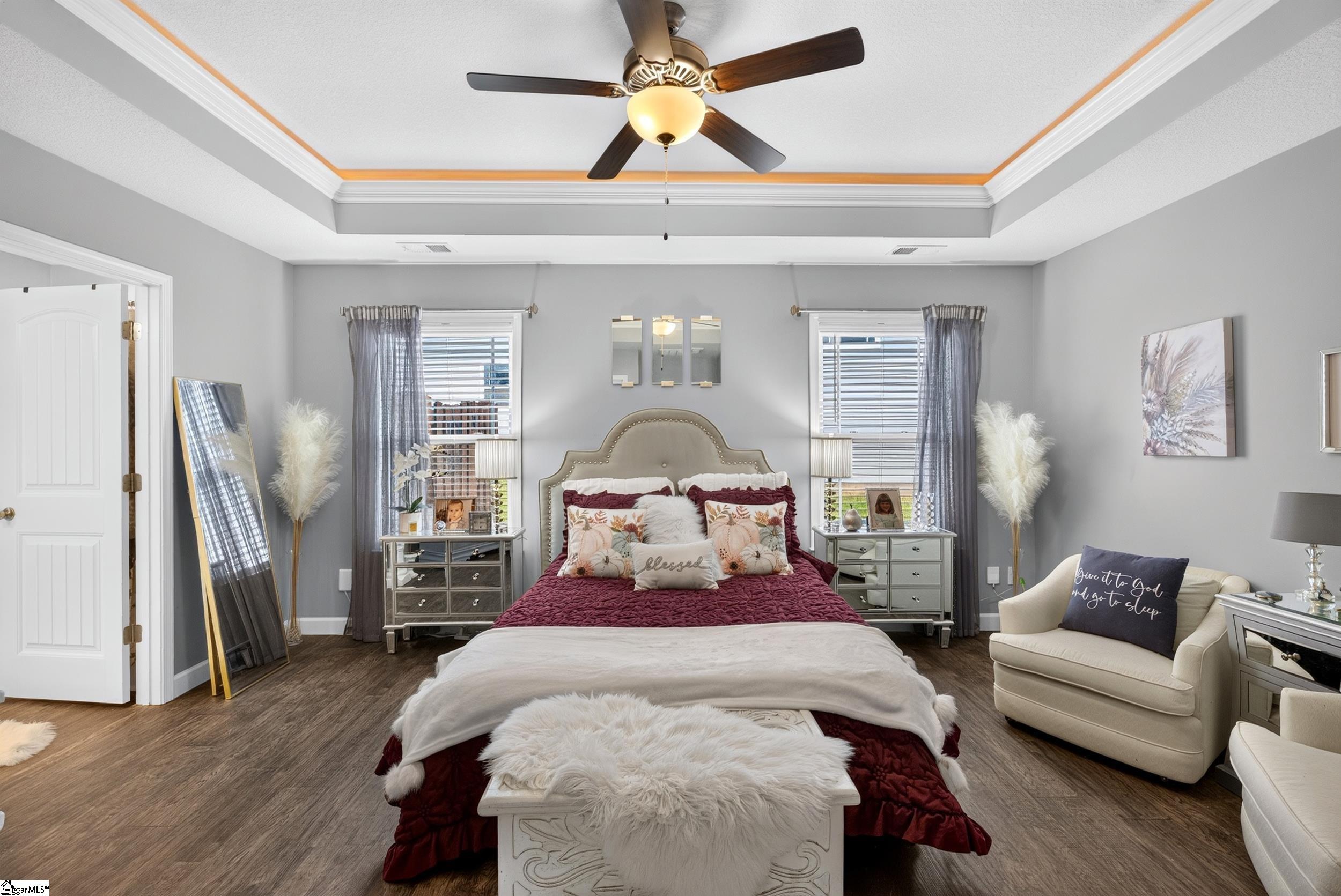 Staged bedroom with a burgundy bed, tray ceiling, ceiling fan, and cream armchair.