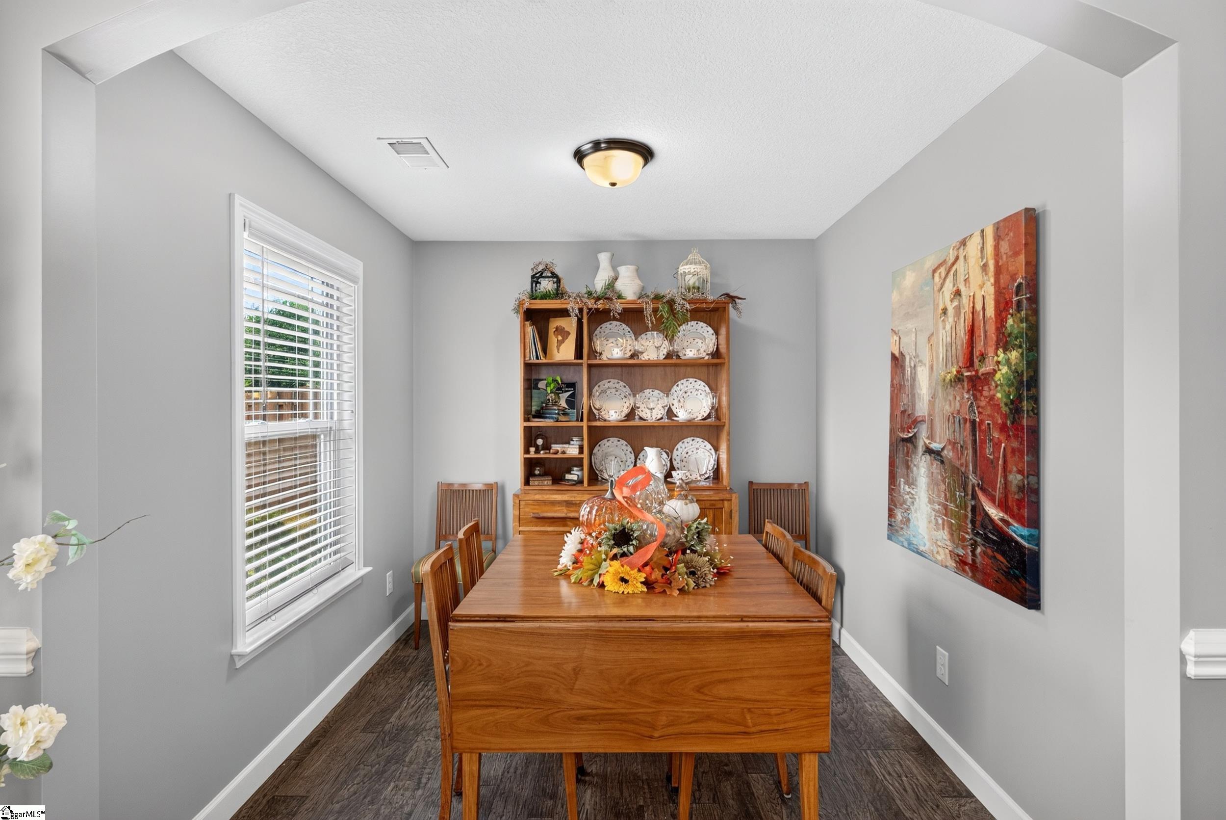 Dining room with a wooden table, china cabinet, window with blinds, and colorful wall art.