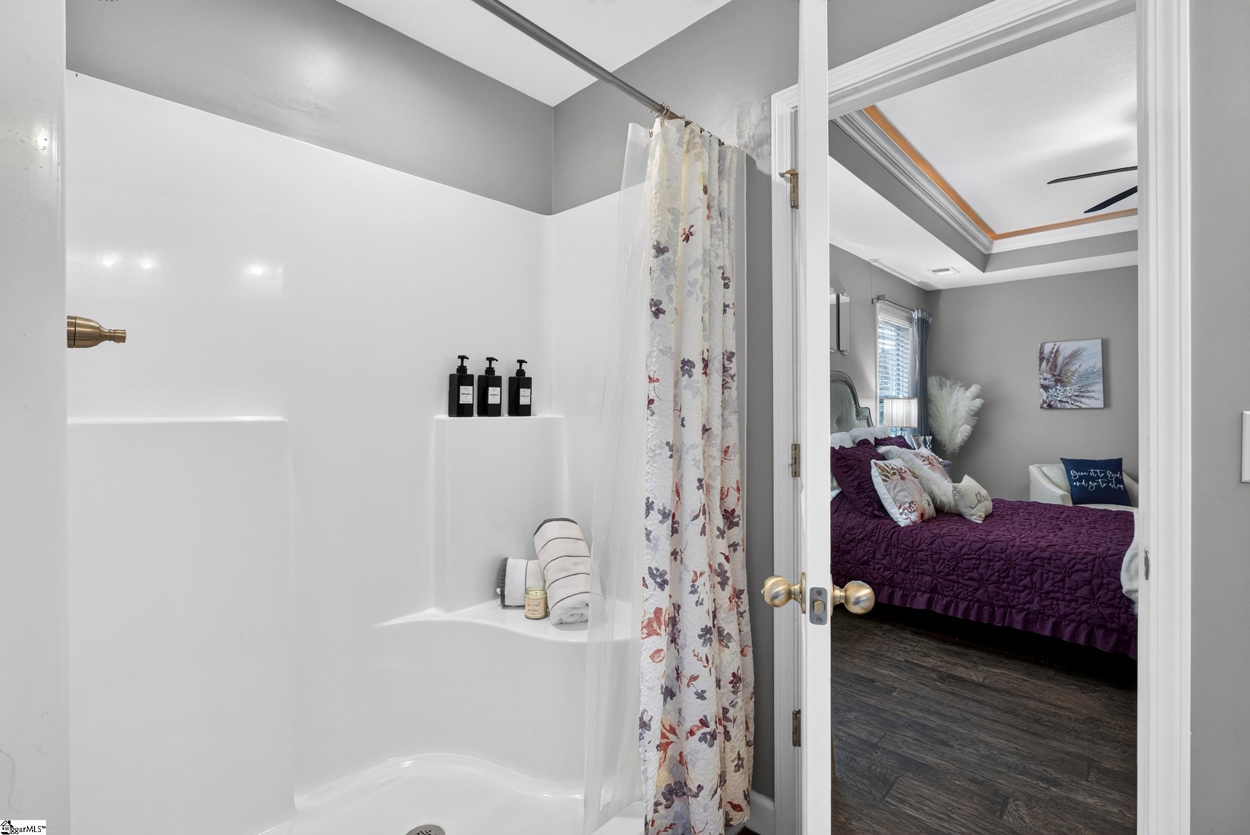 Bathroom with a white tub and floral curtain, opening onto a bedroom with a purple bedspread.