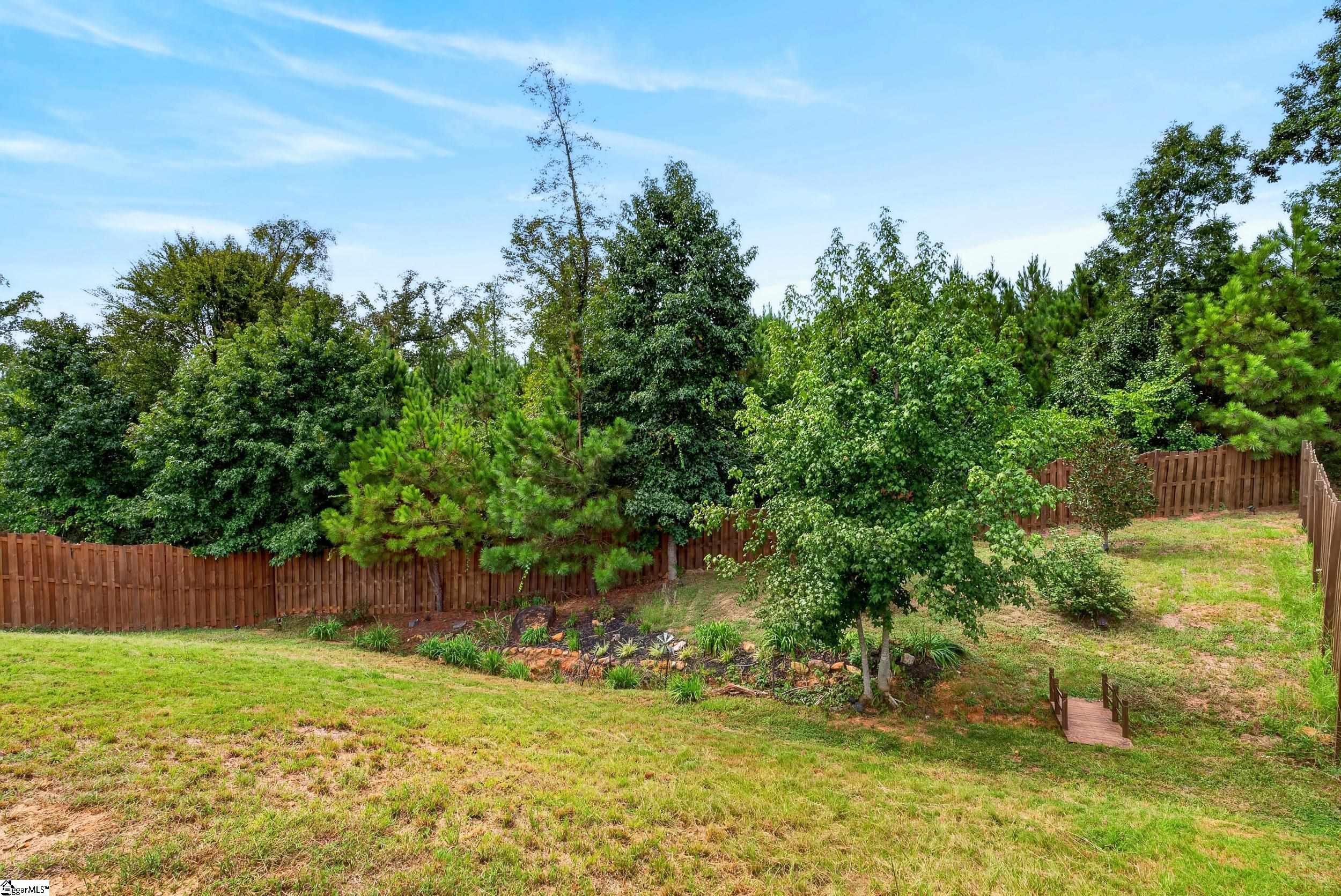 Sloping backyard with a wooden fence, mature trees, landscaped garden bed, and small footbridge.