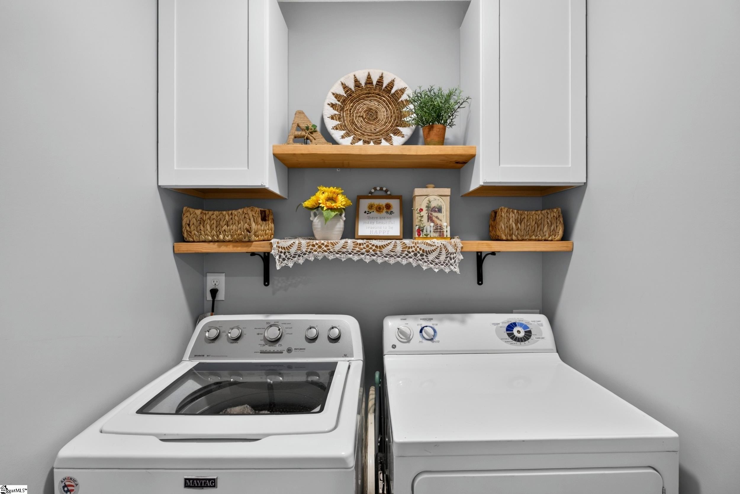 Laundry room with a washer and dryer beneath white cabinets and decorated wooden shelves.