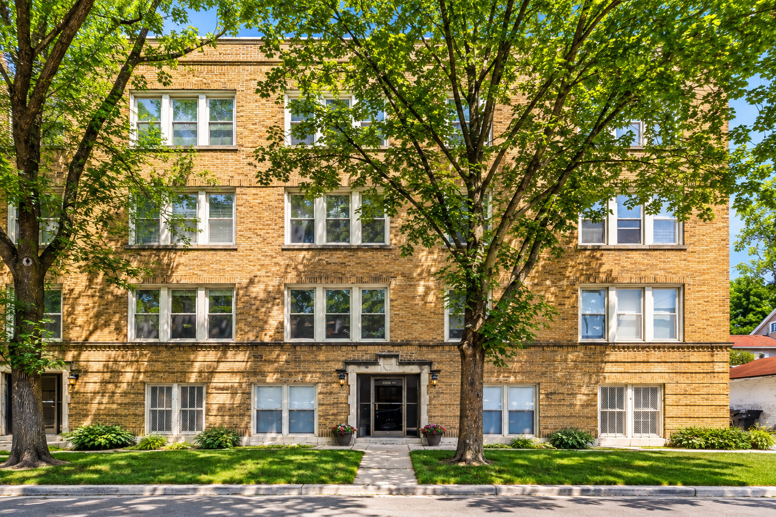 Orange brick apartment building with a central entrance, leafy shade trees, and a front lawn.