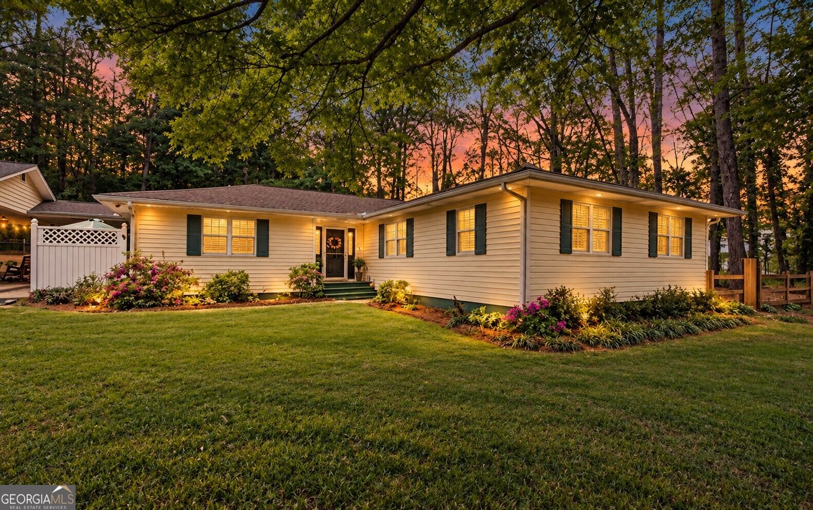 Single-story white home with green shutters and landscaped flower beds at sunset.