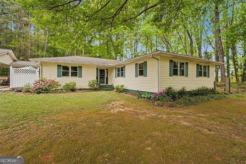 Single-story cream house with green shutters, flowering shrubs, and a wooded lawn.
