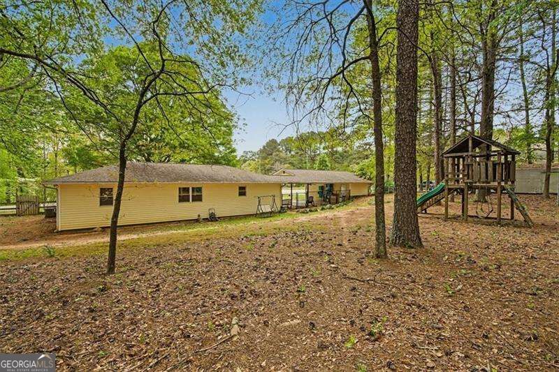 Single-story yellow house in a wooded backyard with a children’s playset and slide.