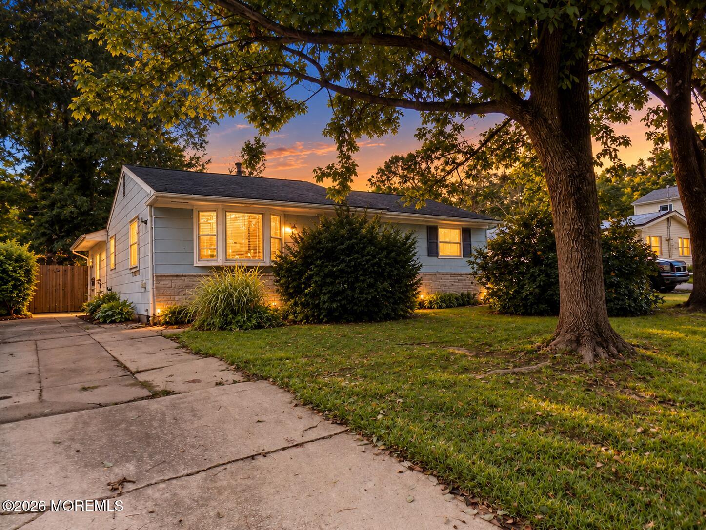Single-story home with blue-gray siding and stone facade beneath a large tree at sunset.