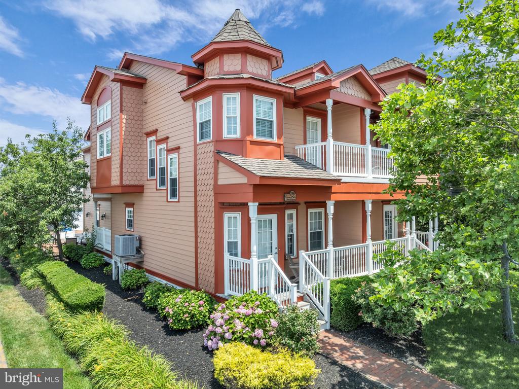 Two-story pink house with a turret, white-railed porches, and flowering garden beds.