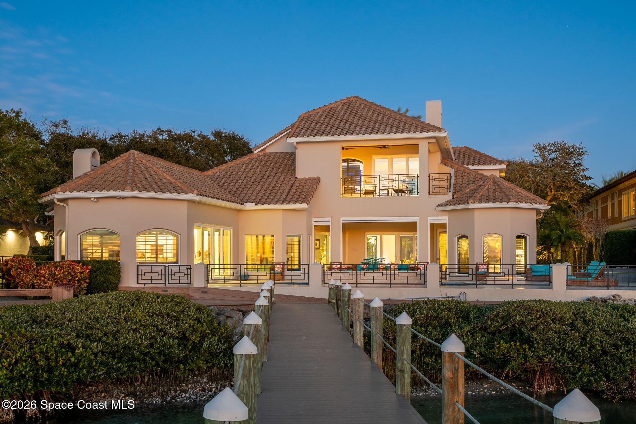 Waterfront home with illuminated windows, a tiled roof, and a boardwalk over the water.