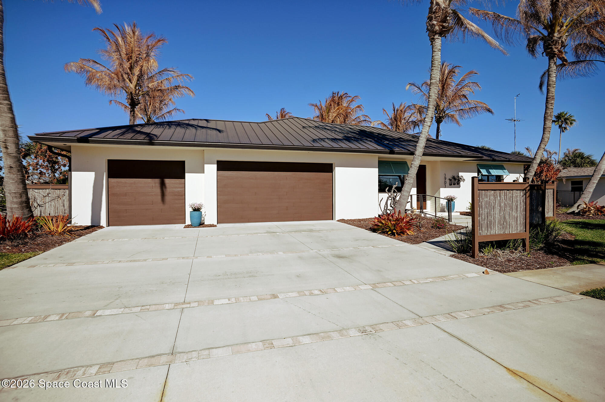 Modern single-story home with a dark metal roof, double garage, broad driveway, and palm trees.