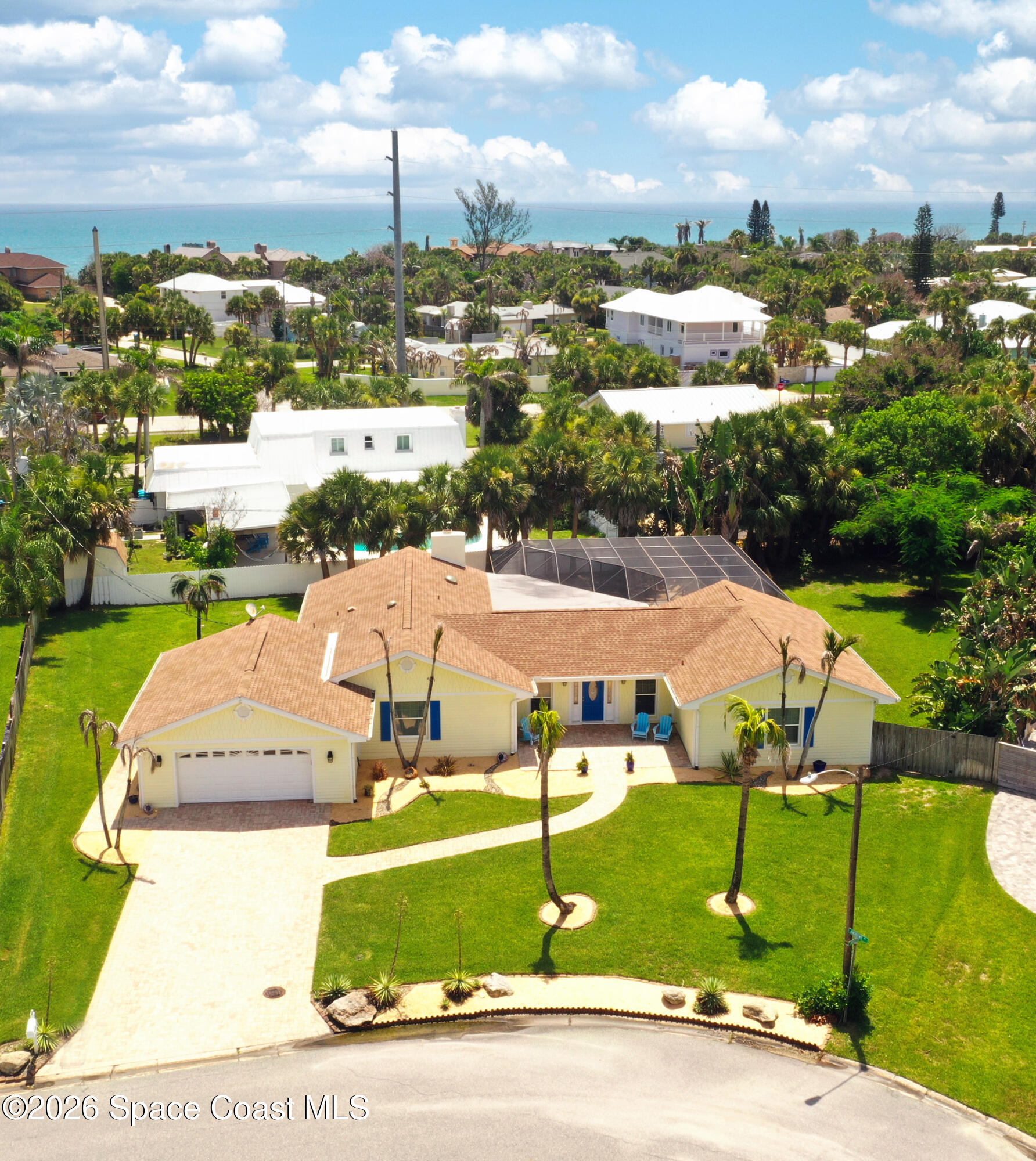 Aerial view of a yellow coastal home with a curved driveway, palm trees, screened pool enclosure, and ocean beyond.