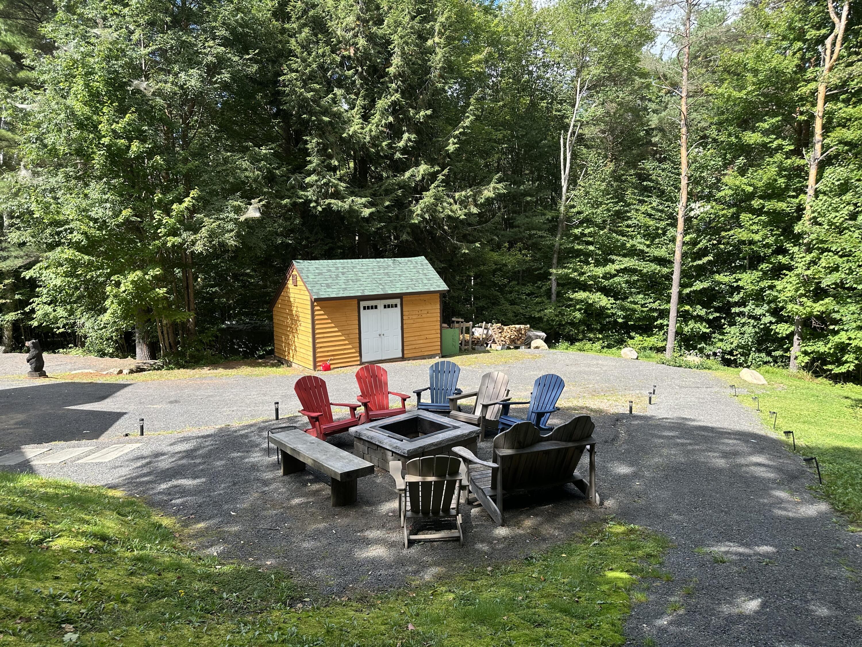 Gravel patio with a stone fire pit, Adirondack chairs, and a wooden shed amid trees.