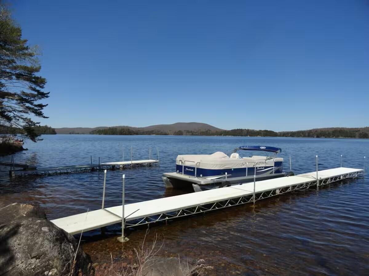 White dock and pontoon boat on a lake, with rocky shore and wooded hills beyond.