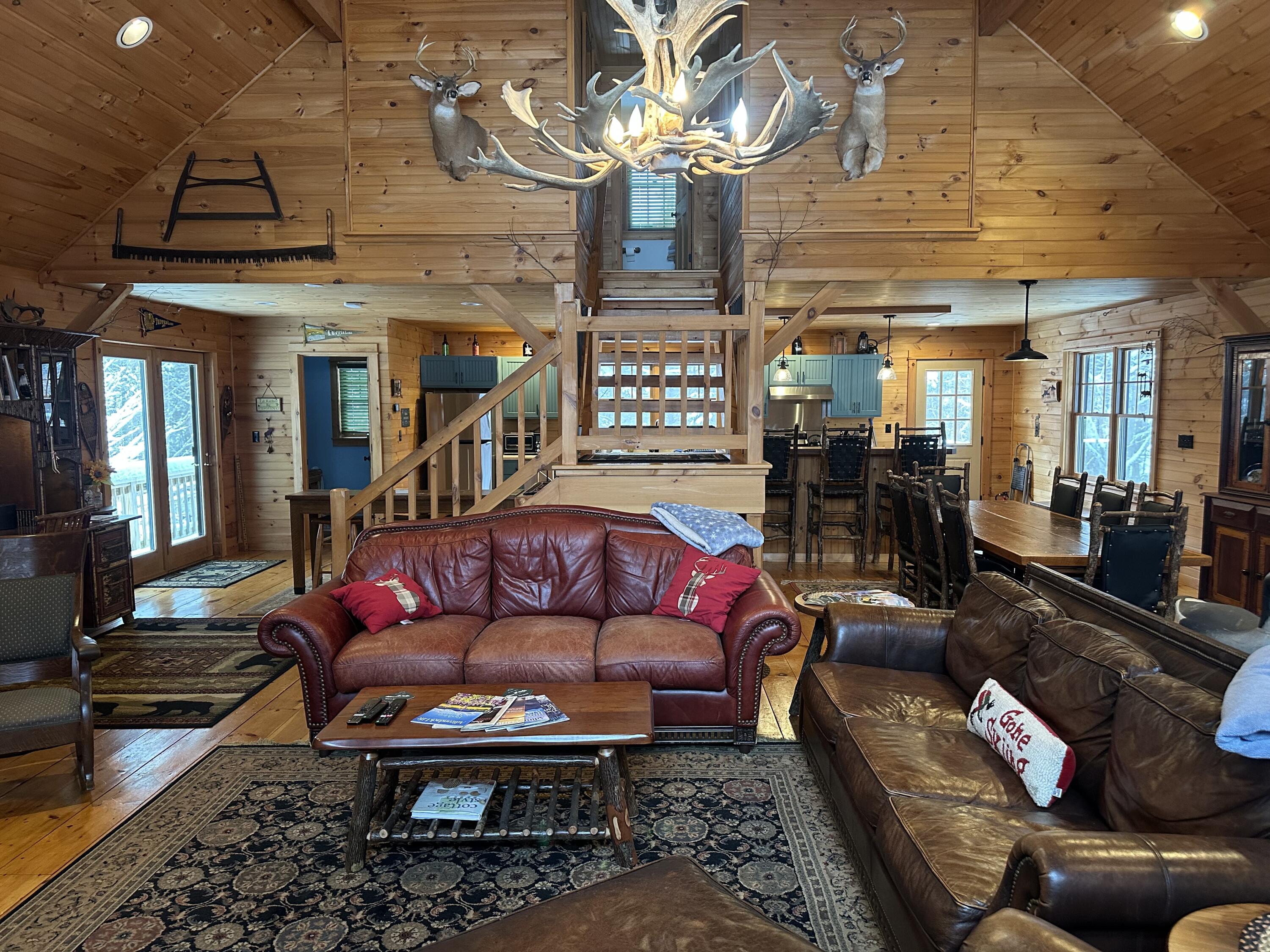 Rustic wood-paneled living room with leather sofas, an antler chandelier, and stairs to a loft.