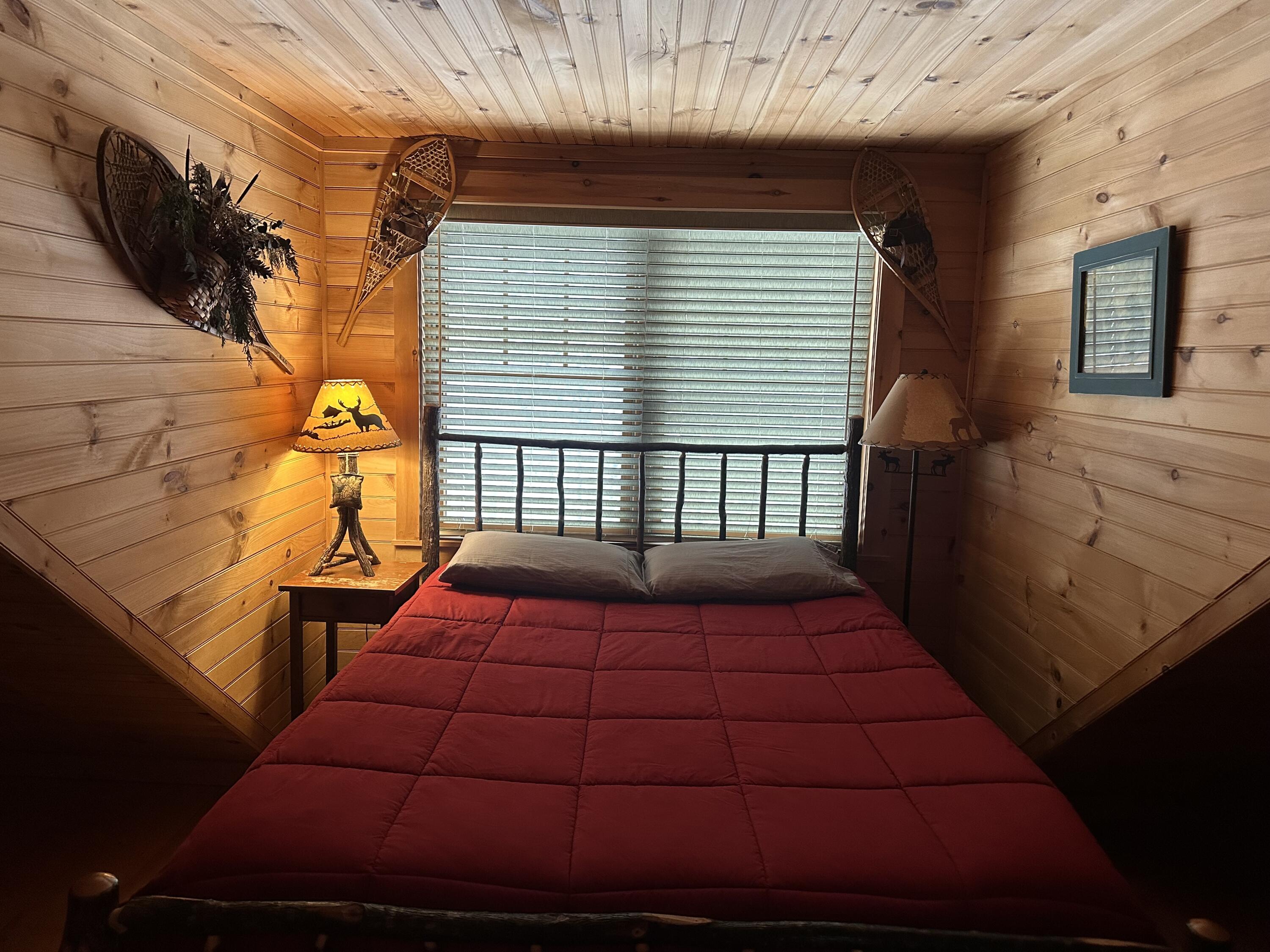 Bedroom with a red quilted bed, wood-paneled walls, and rustic snowshoes above the window.