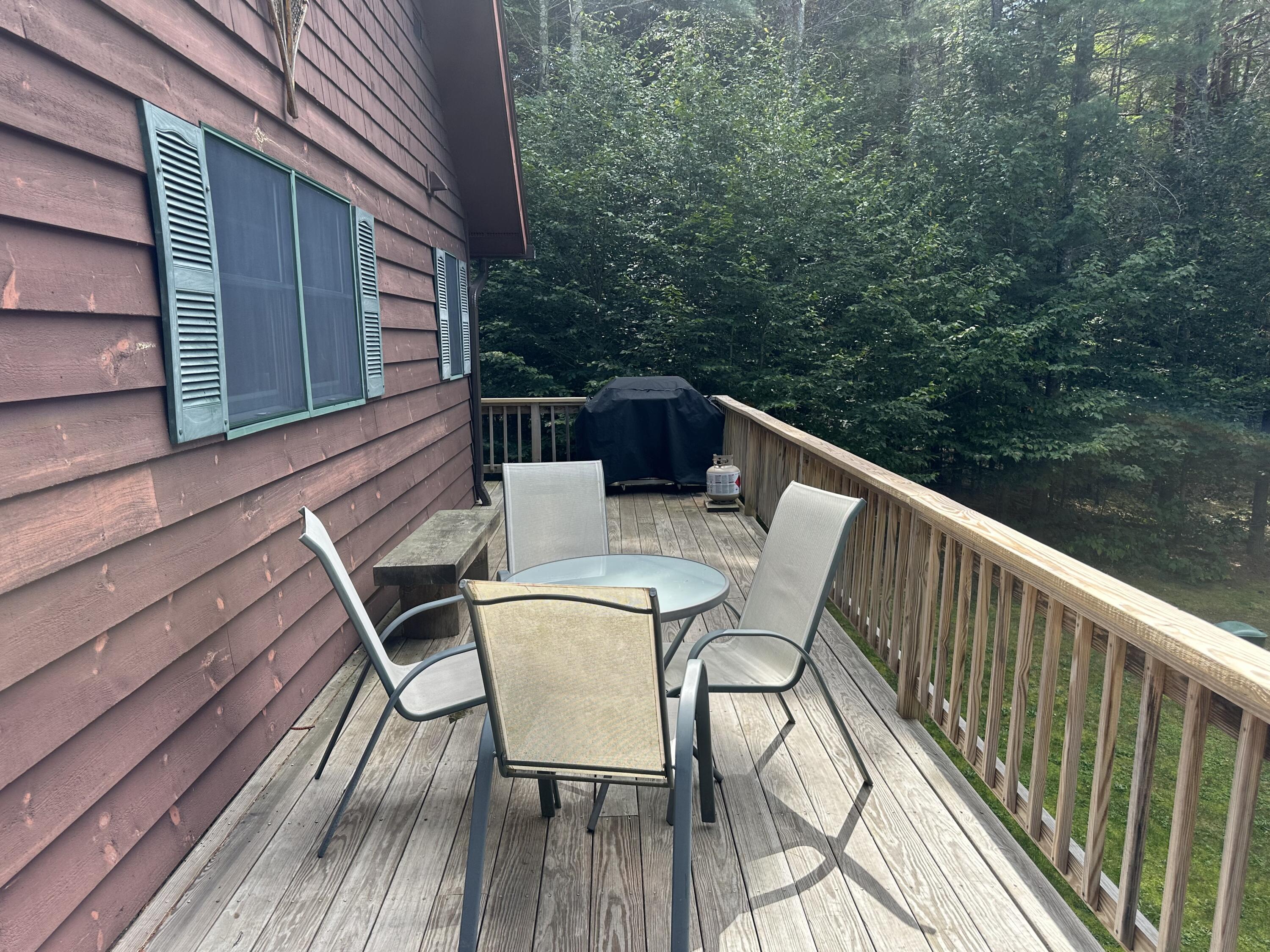 Wooden deck with patio table and chairs beside a brown-sided house, surrounded by trees.