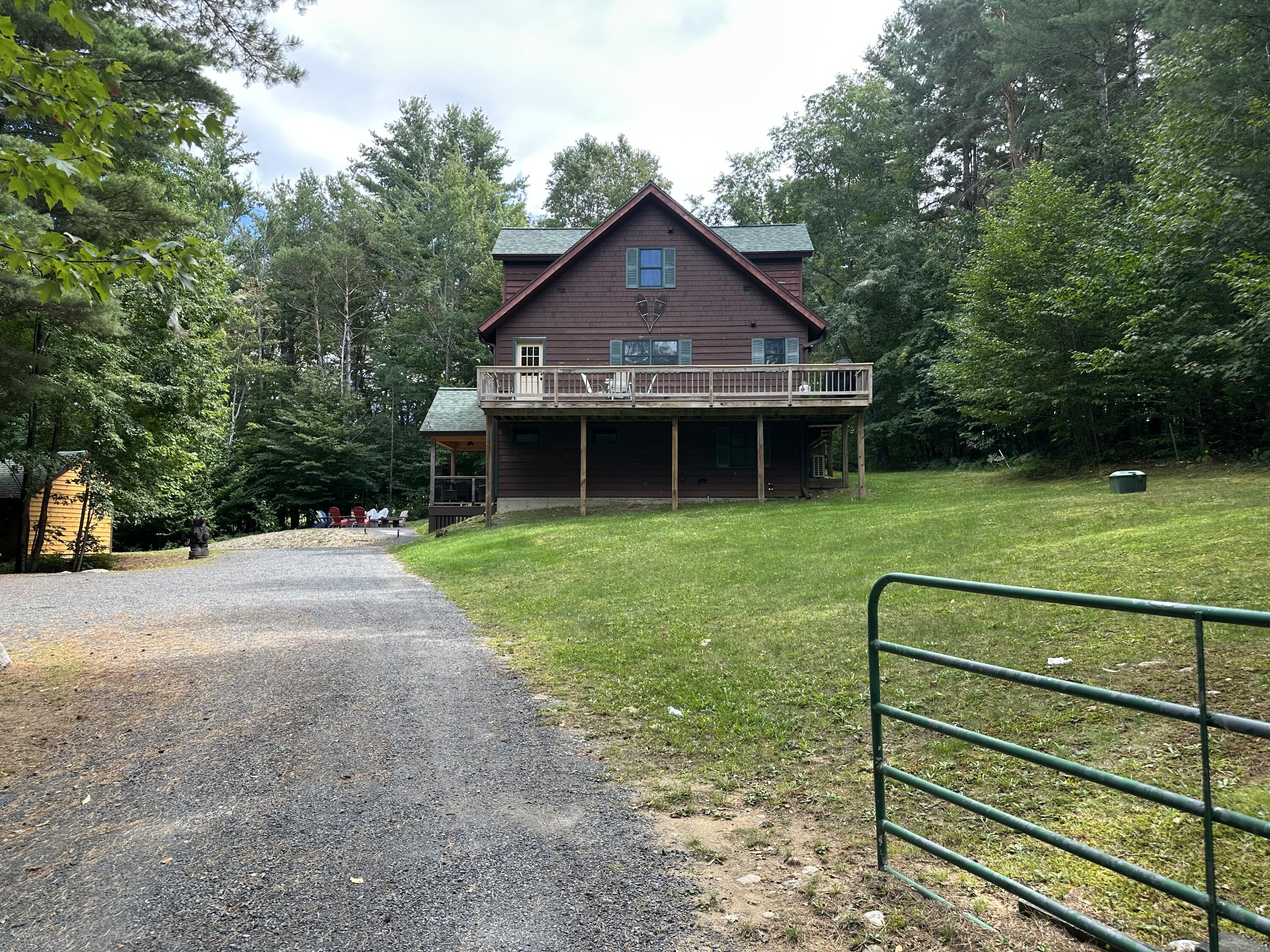 Dark brown house with a broad deck among trees, viewed from the gravel driveway and lawn.