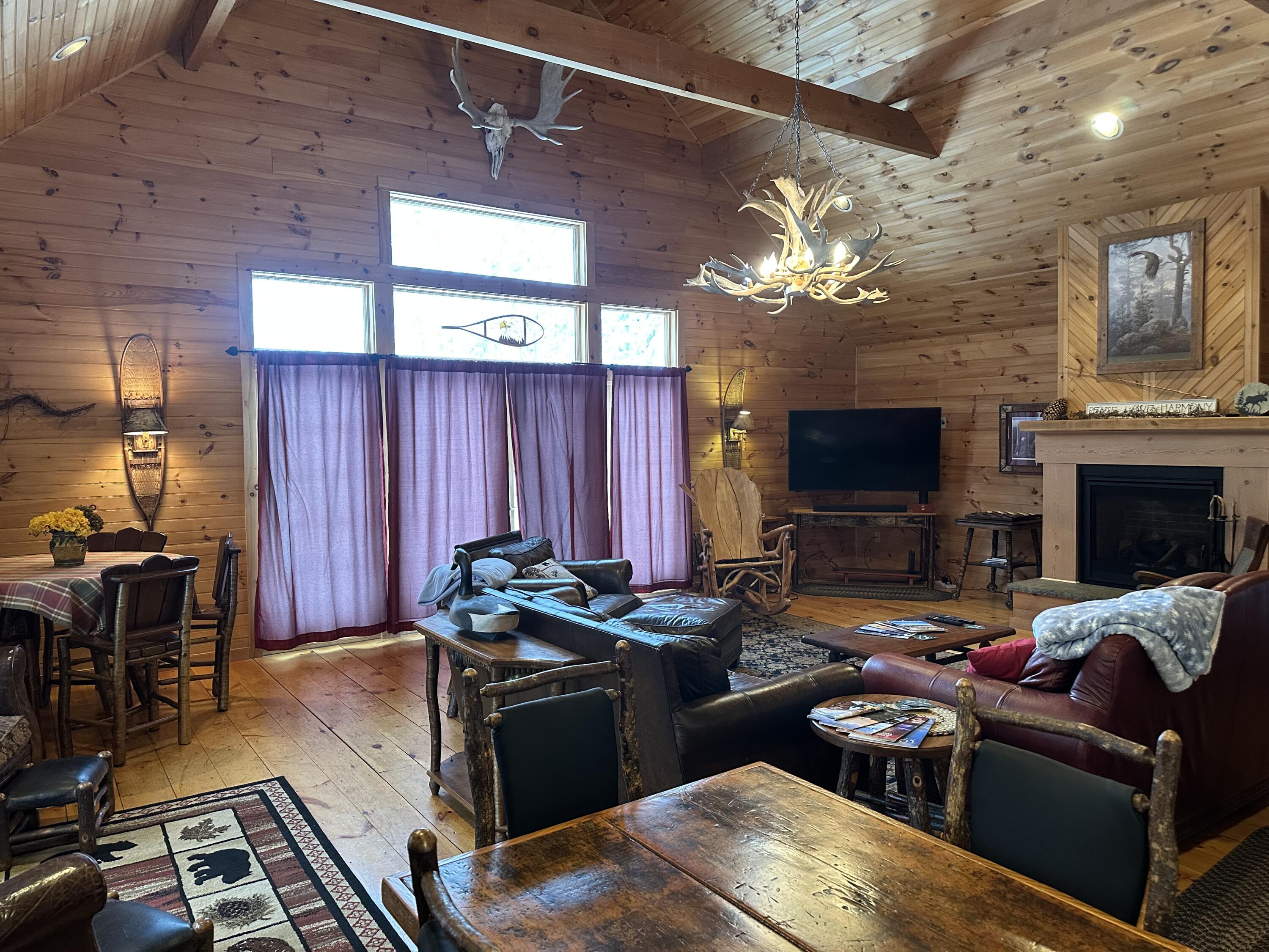 Wood-paneled living room with vaulted ceiling, antler chandelier, leather seating, and fireplace.