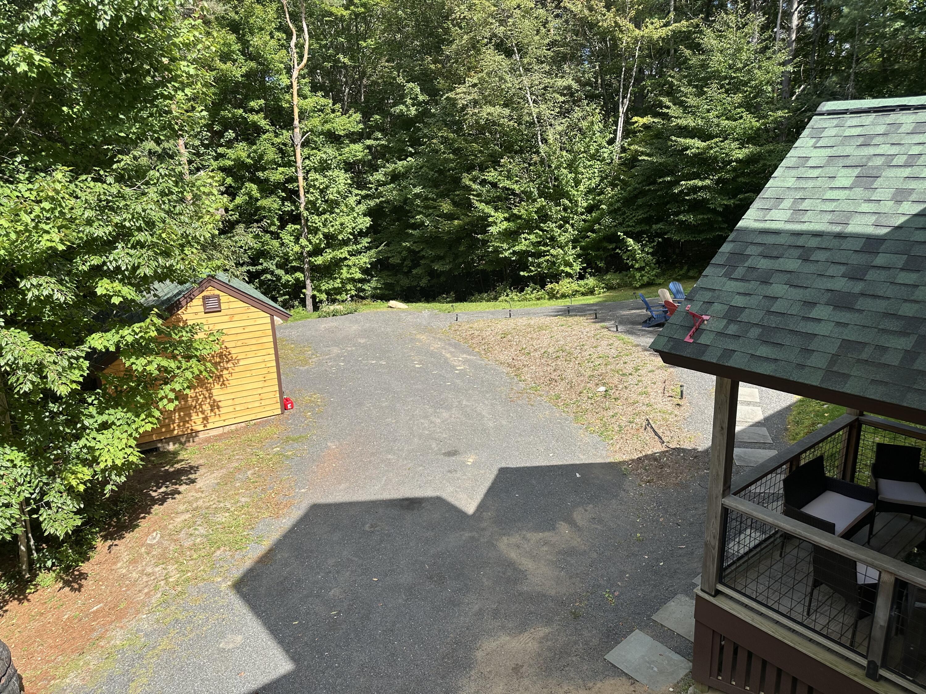 Gravel driveway and wooden shed beside a covered deck, surrounded by dense summer woods.