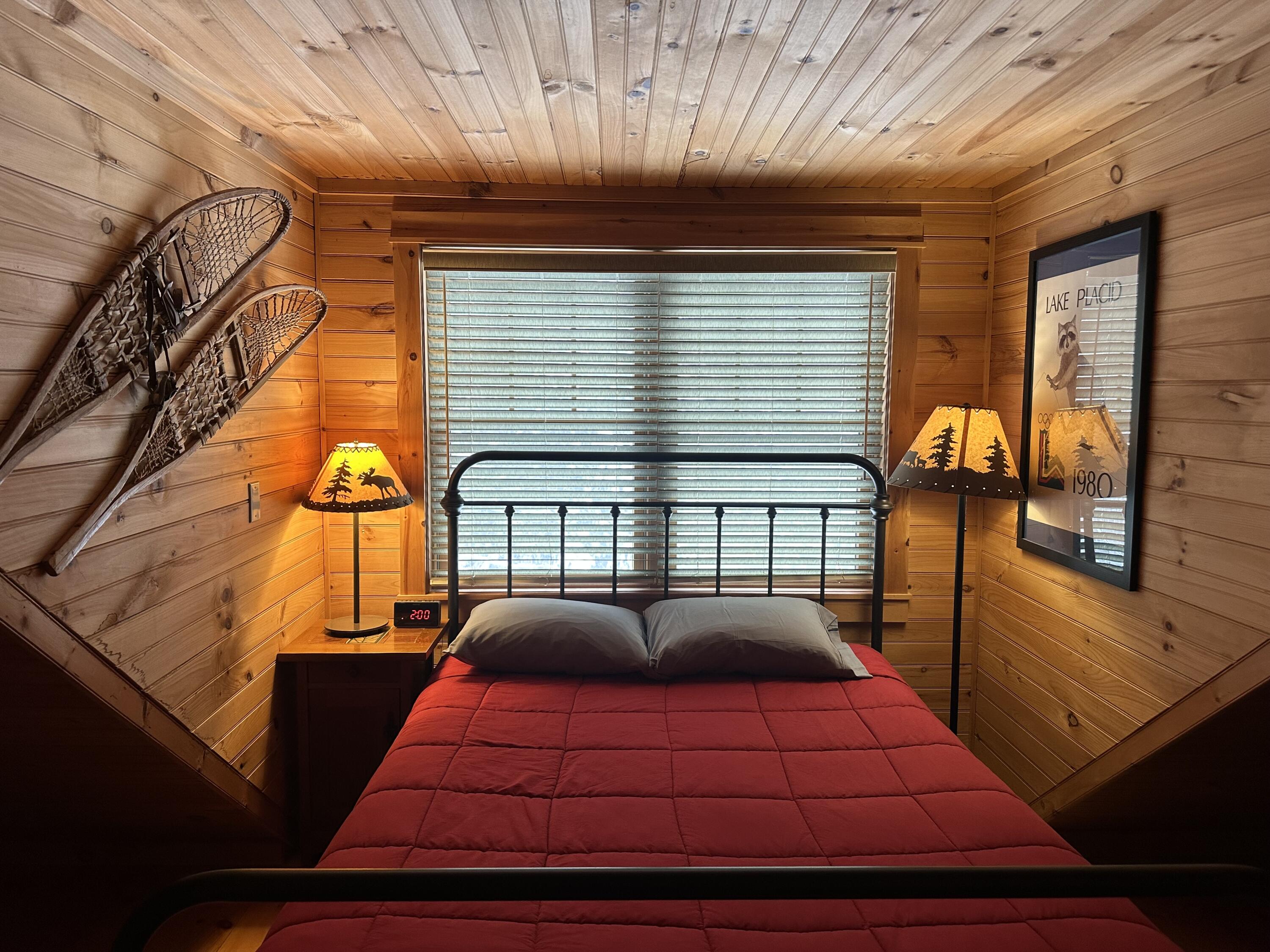Wood-paneled bedroom with a red bed, wall-mounted snowshoes, and rustic Adirondack lamps.