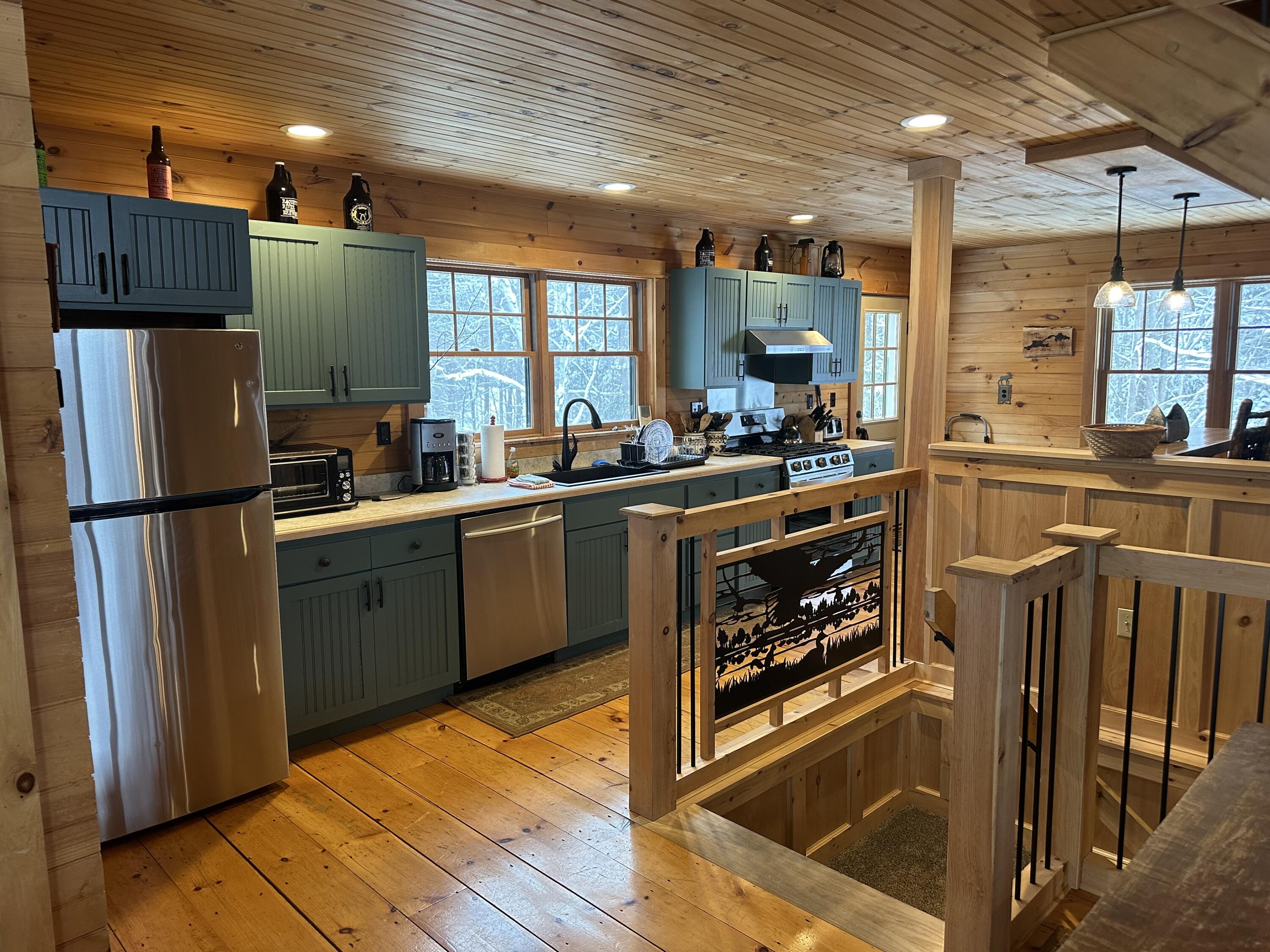 Rustic wood-paneled kitchen with green cabinets and stainless steel appliances, beside a stairwell railing.