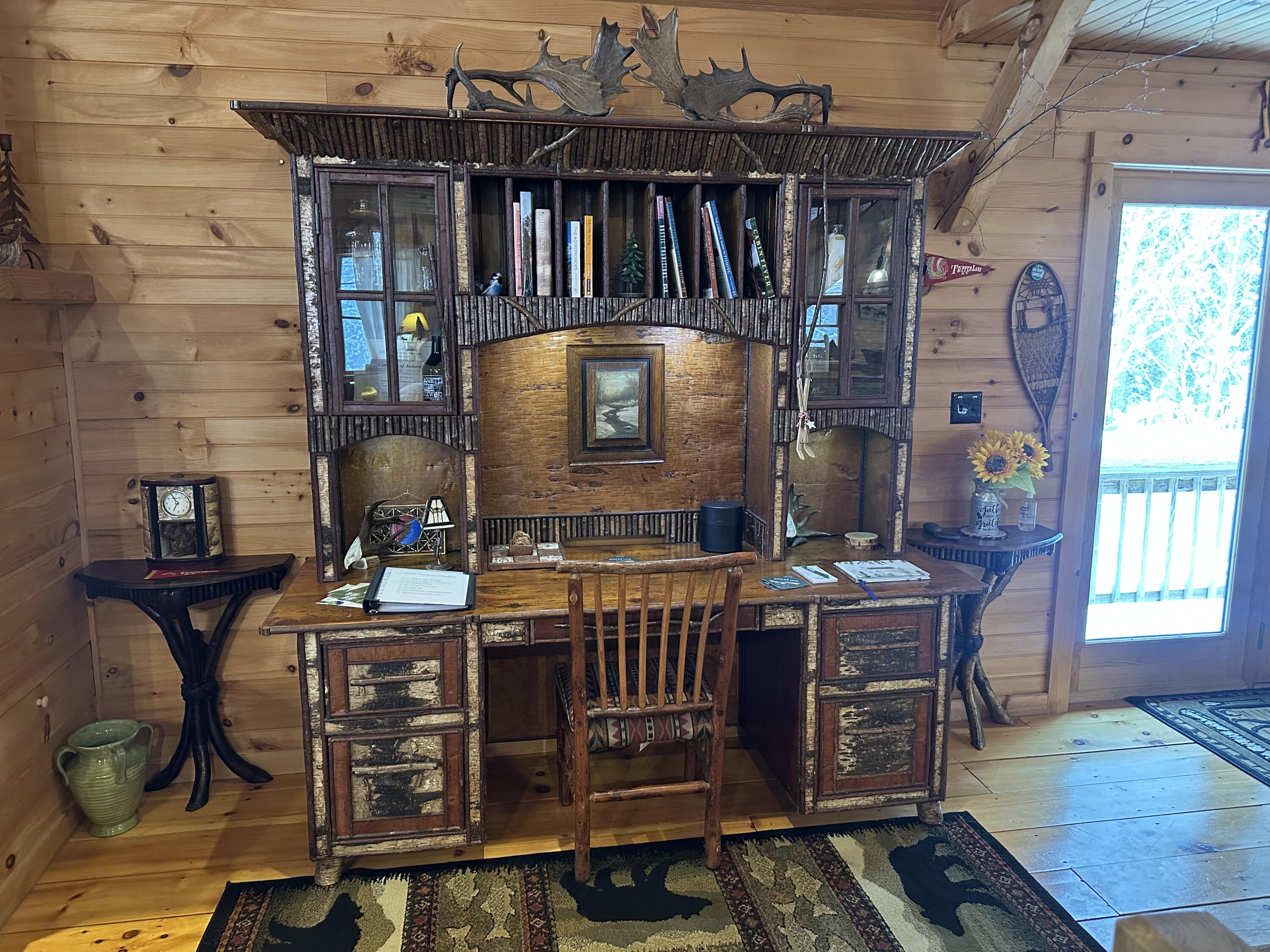 Rustic cabin study with an ornate desk, bookcase hutch, antler decor, and a bright deck doorway.