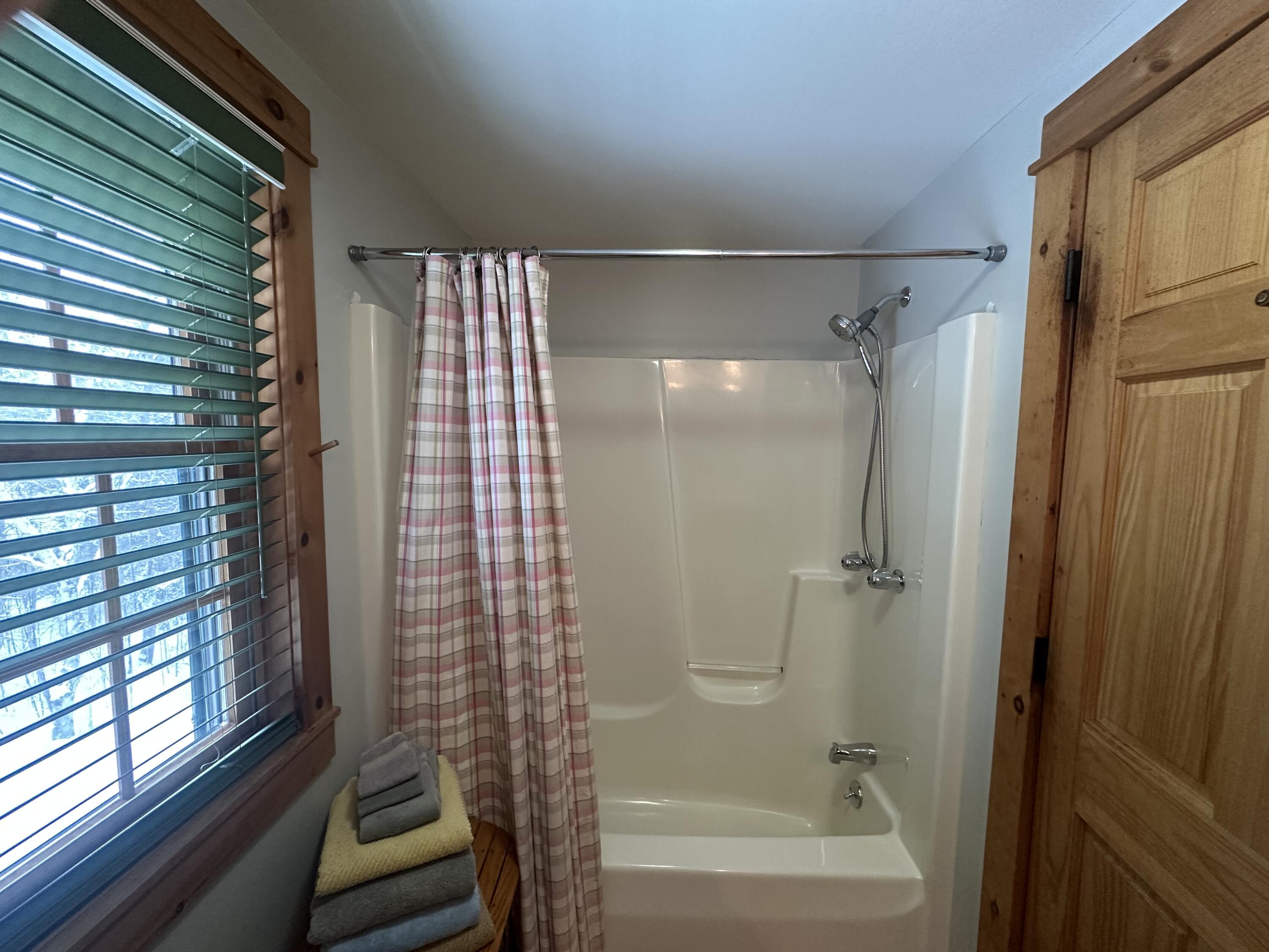 Bathroom tub and shower with a plaid curtain beside a wood-trimmed window and paneled door.