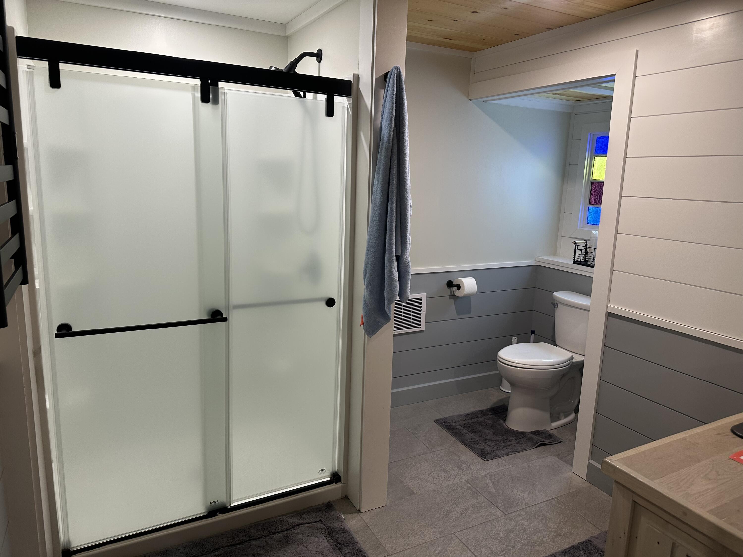 Bathroom with frosted-glass sliding shower doors, gray tile, and a toilet beneath a small stained-glass window.