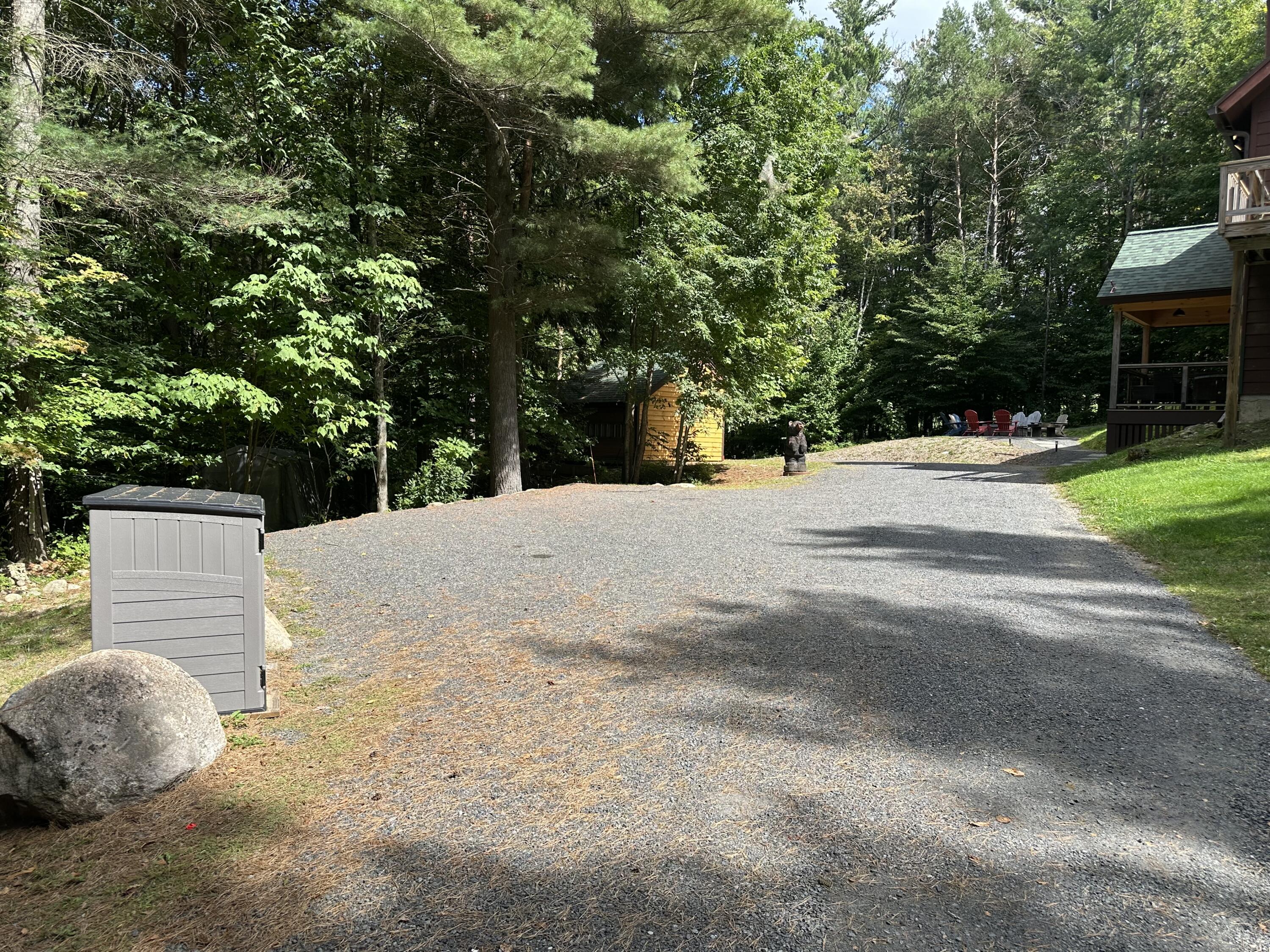 Gravel driveway through a wooded property, with cabins, outdoor chairs, and tall trees.