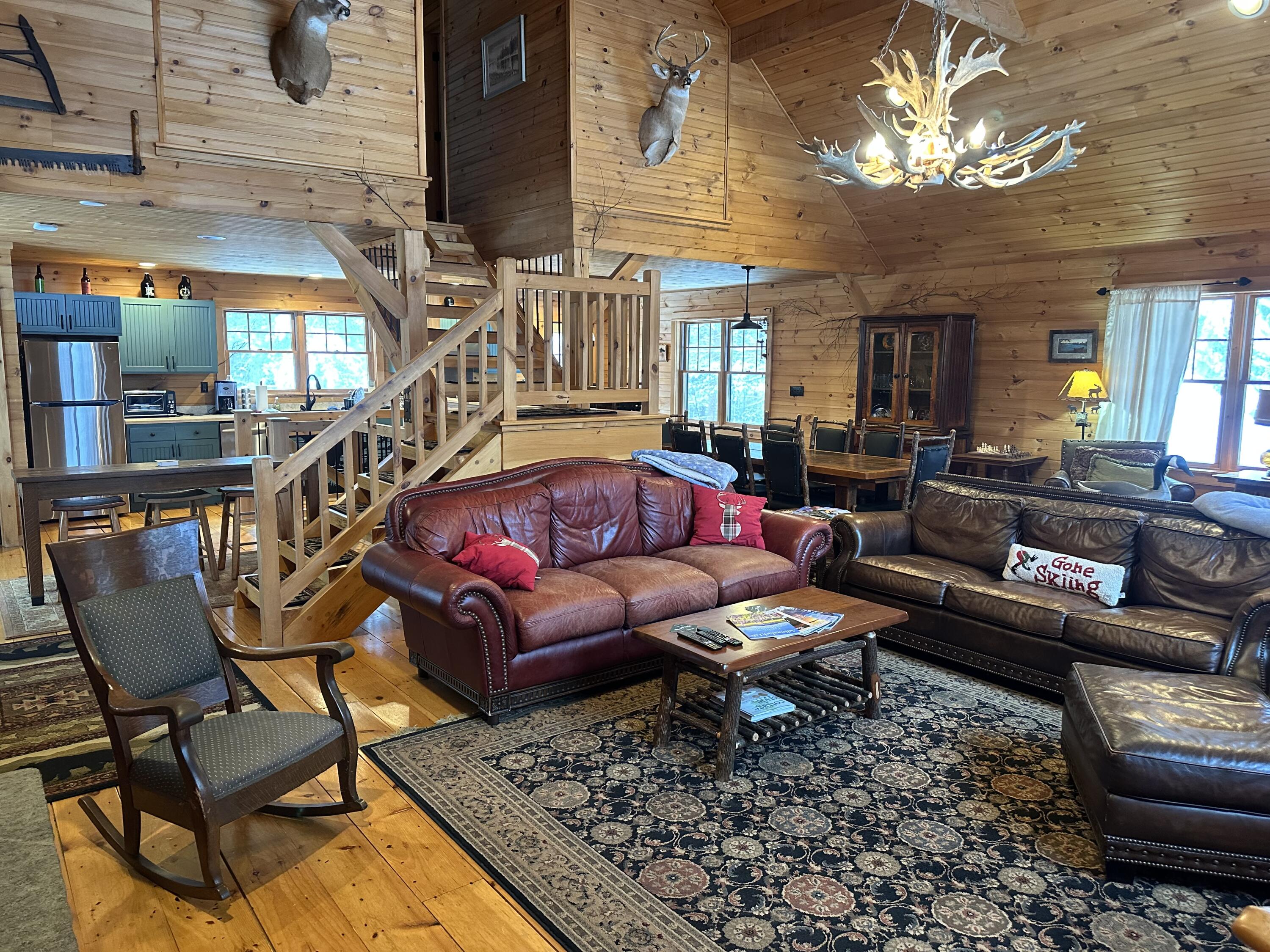 Rustic living room with leather sofas, a central staircase, and an open kitchen beneath wood-paneled ceilings.