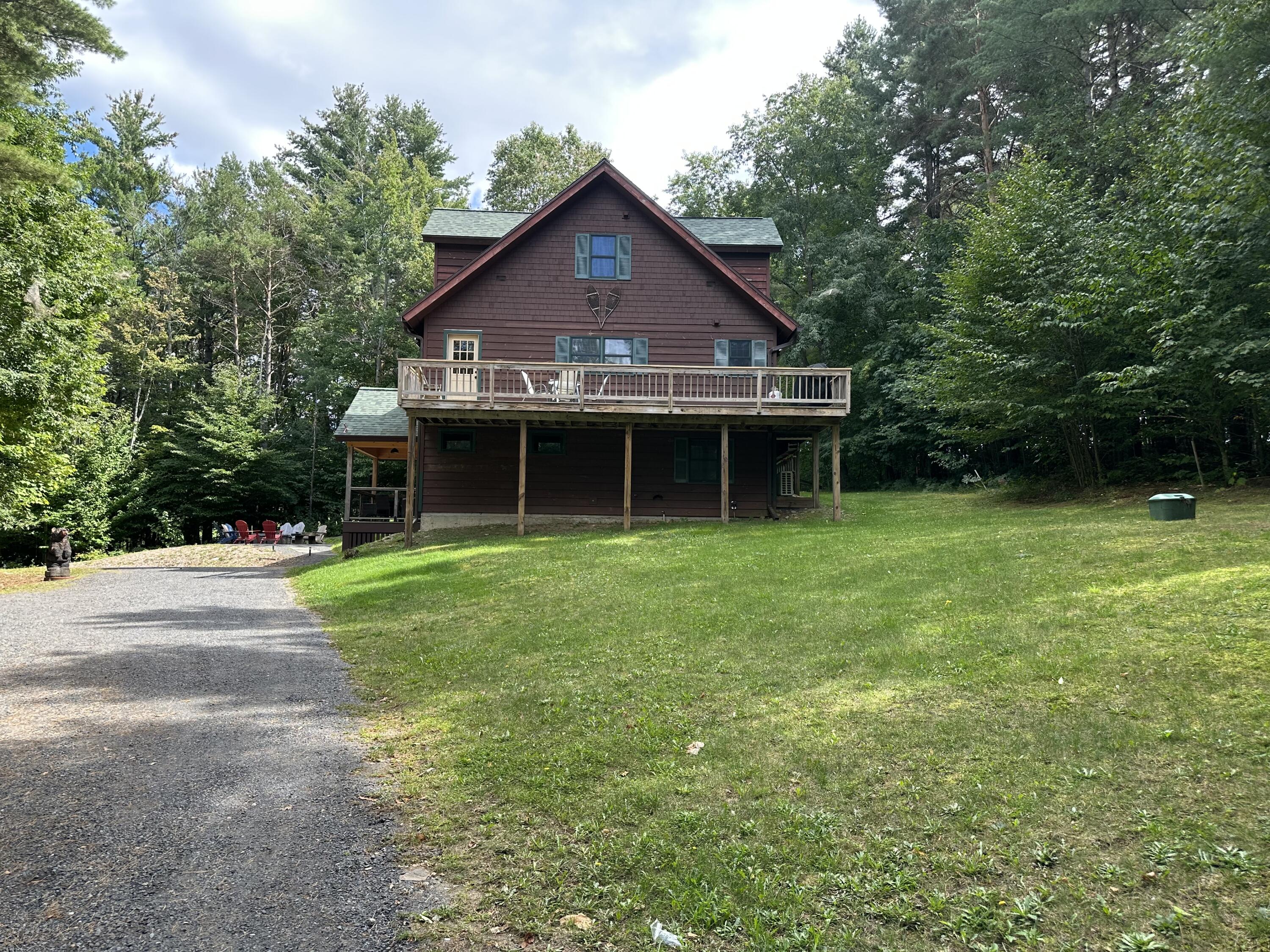 Brown multi-level house with a raised deck, surrounded by trees beside a gravel driveway.