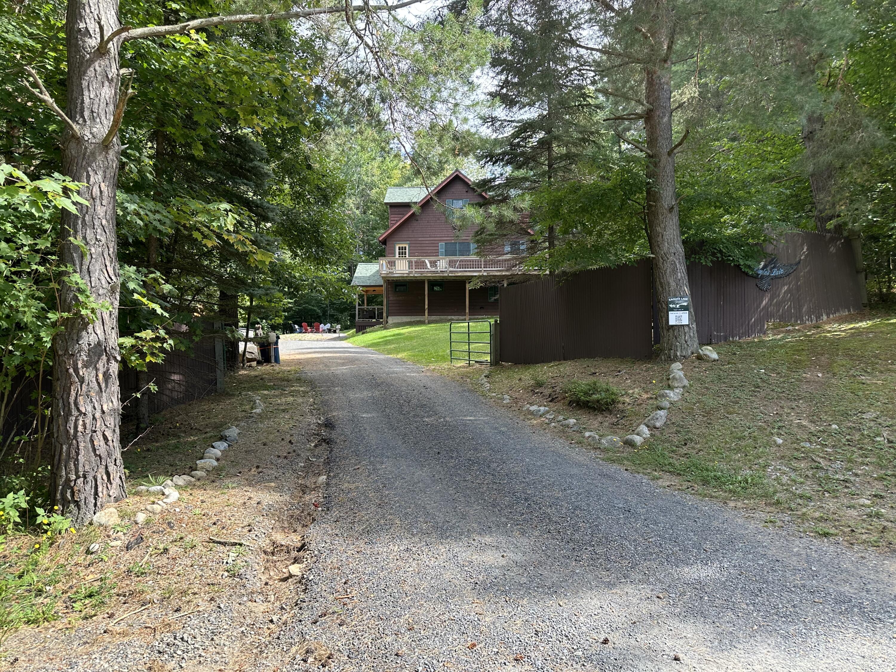 Wooded driveway leading to a red-brown house with a balcony and fenced entrance.