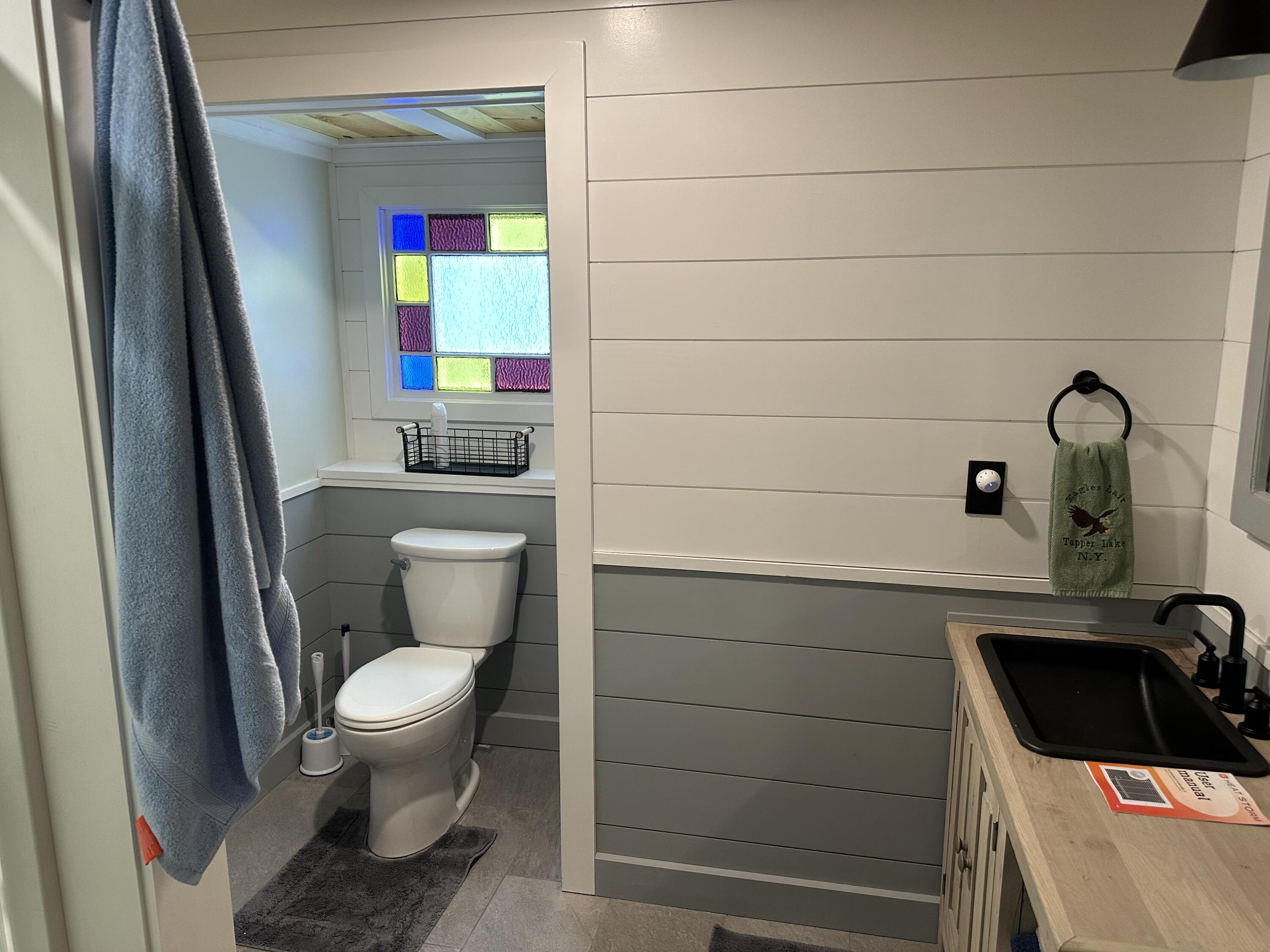 Bathroom with a colorful stained-glass window, gray paneled walls, and a wood vanity with a black sink.