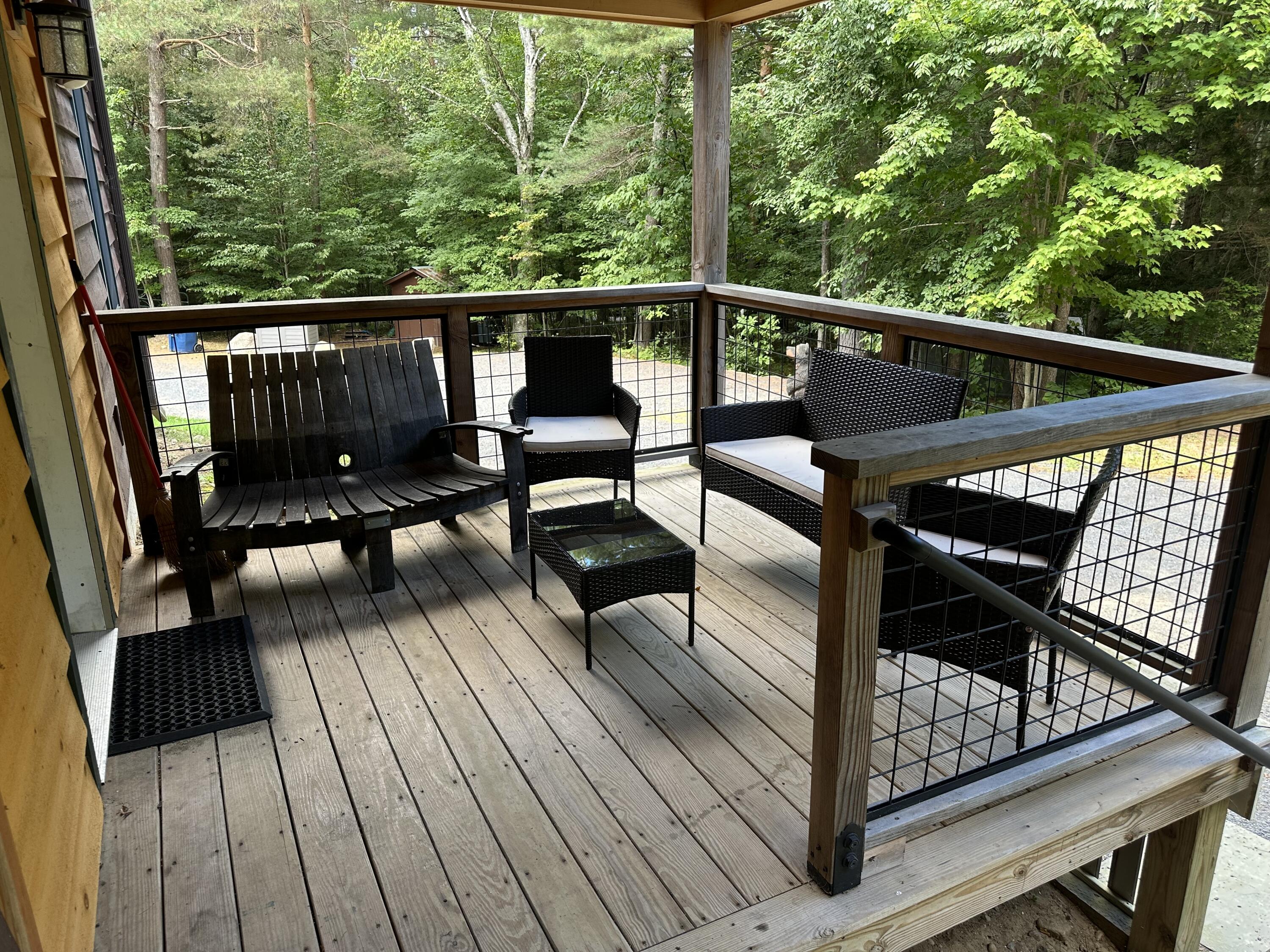 Furnished wooden deck overlooking trees, with a wooden bench, wicker chairs, and black grid railings.