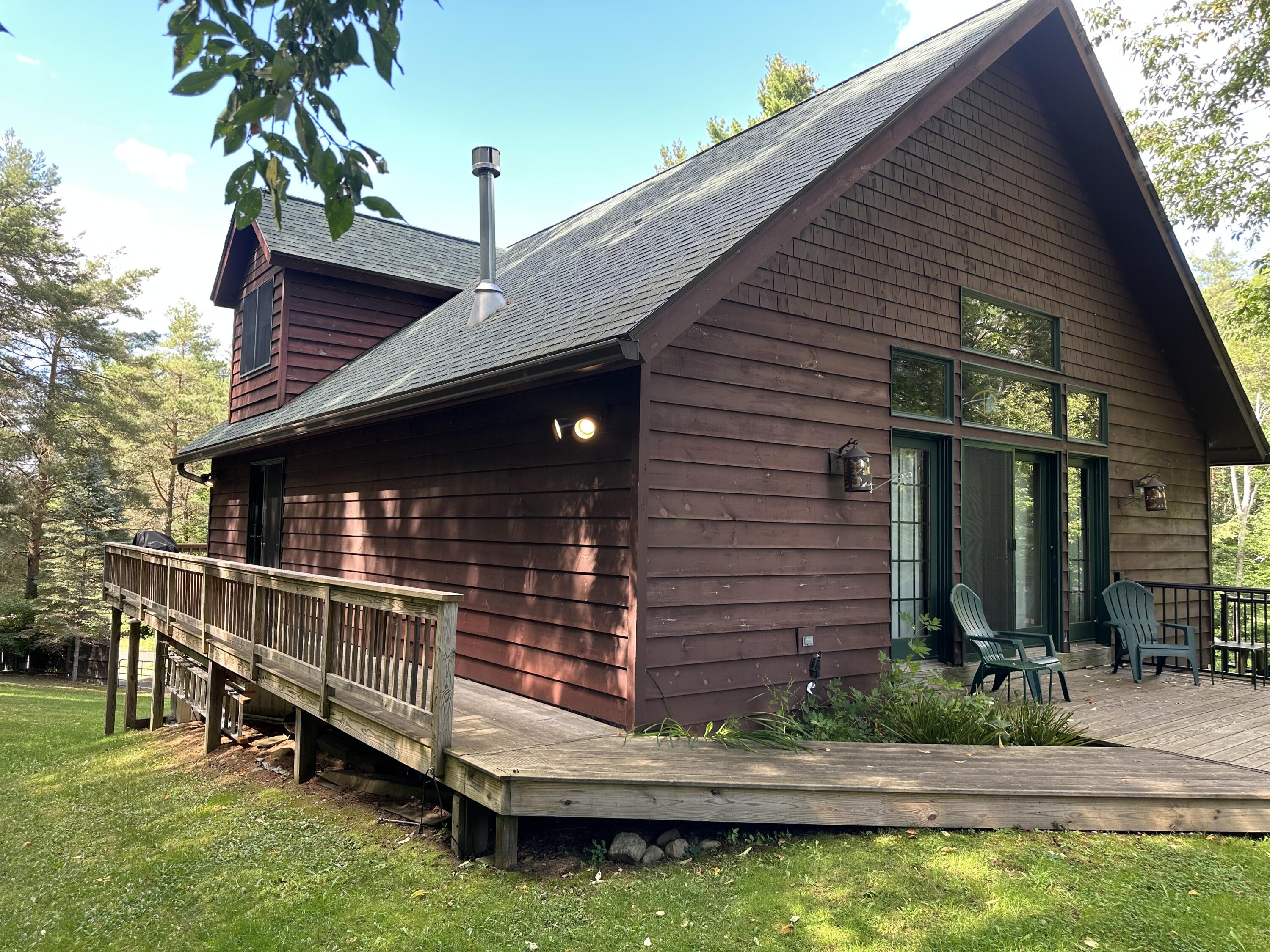 Wood-sided A-frame home with a wraparound deck and patio chairs among trees.