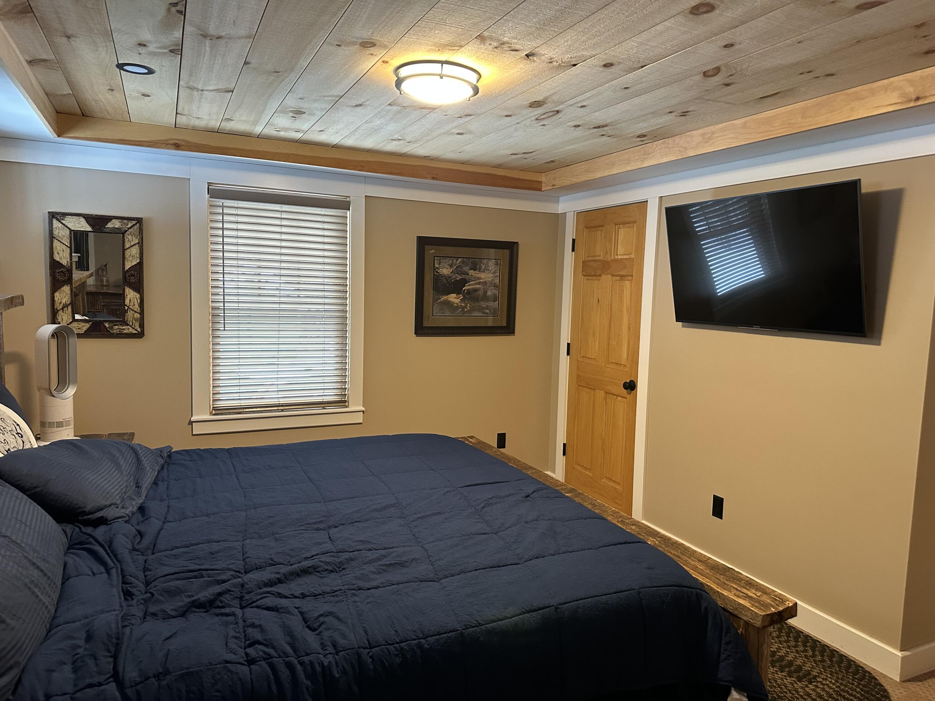 Bedroom with a large dark blue bed, wood-plank ceiling, and wall-mounted television.