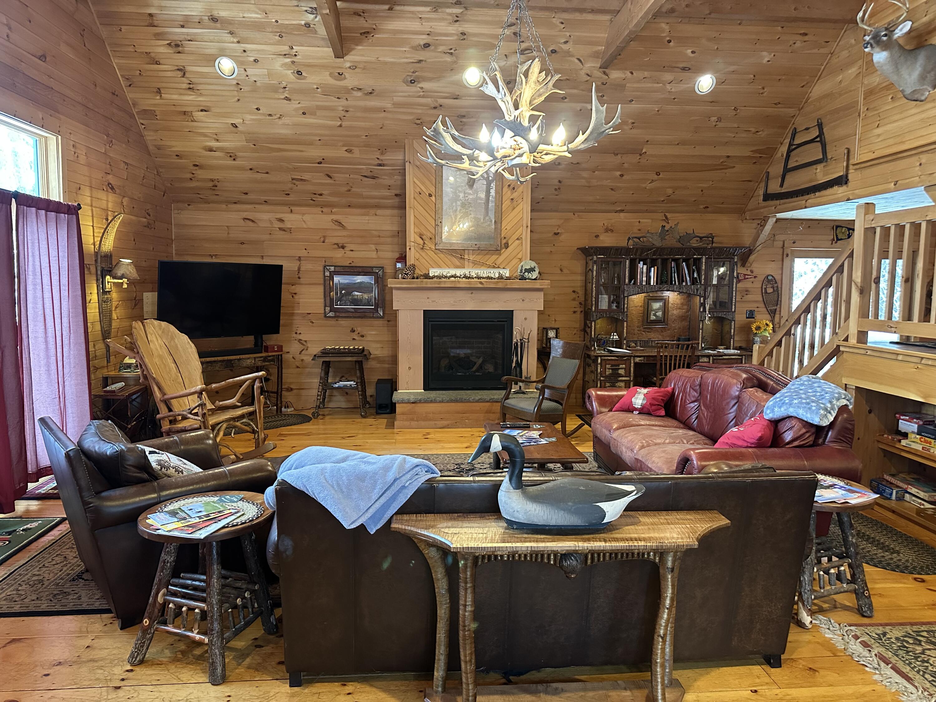 Rustic living room with a central fireplace, leather seating, wood-paneled ceiling, and antler chandelier.