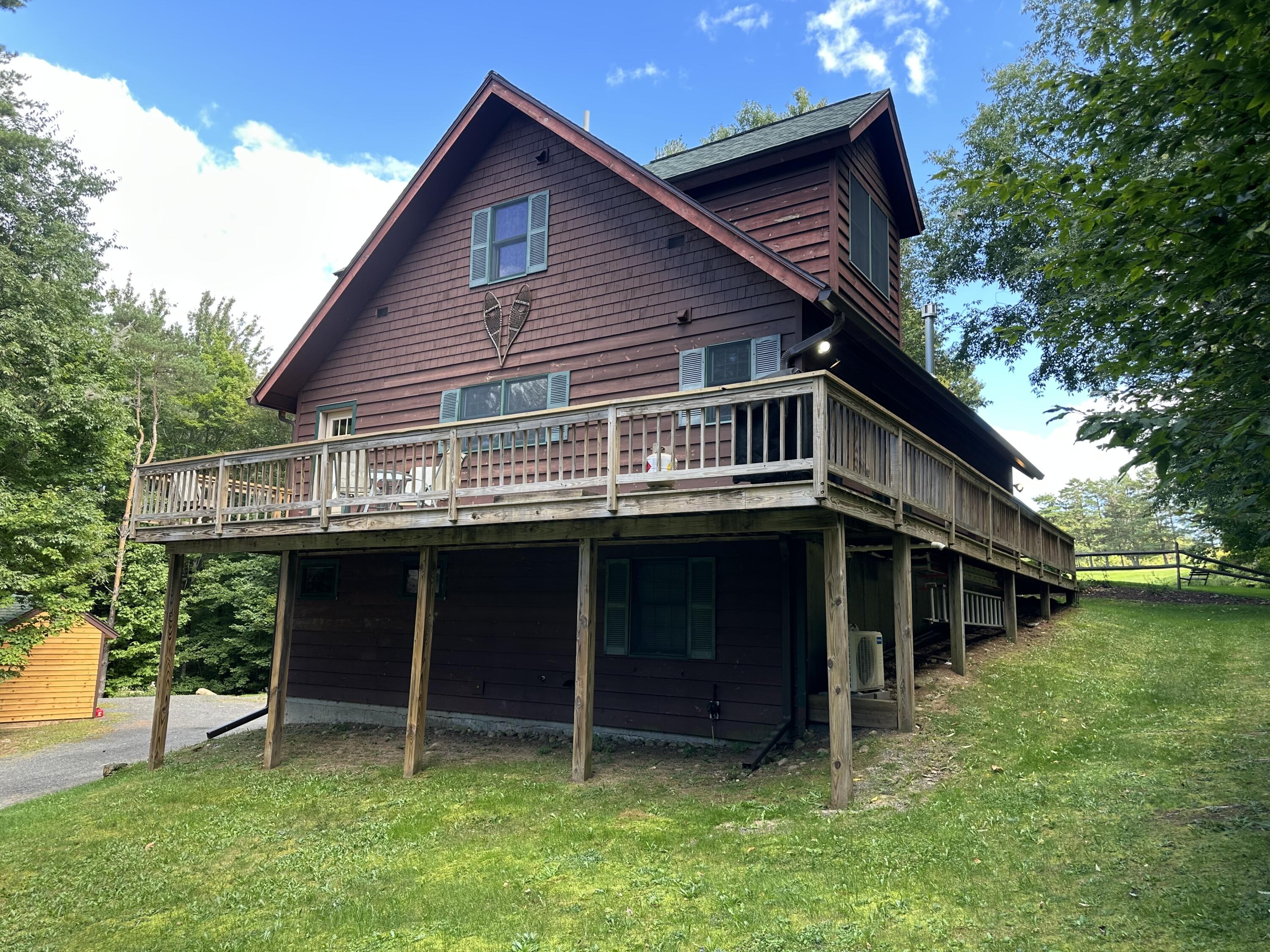 Brown multi-level home with a large elevated wooden deck, surrounded by trees and a grassy yard.