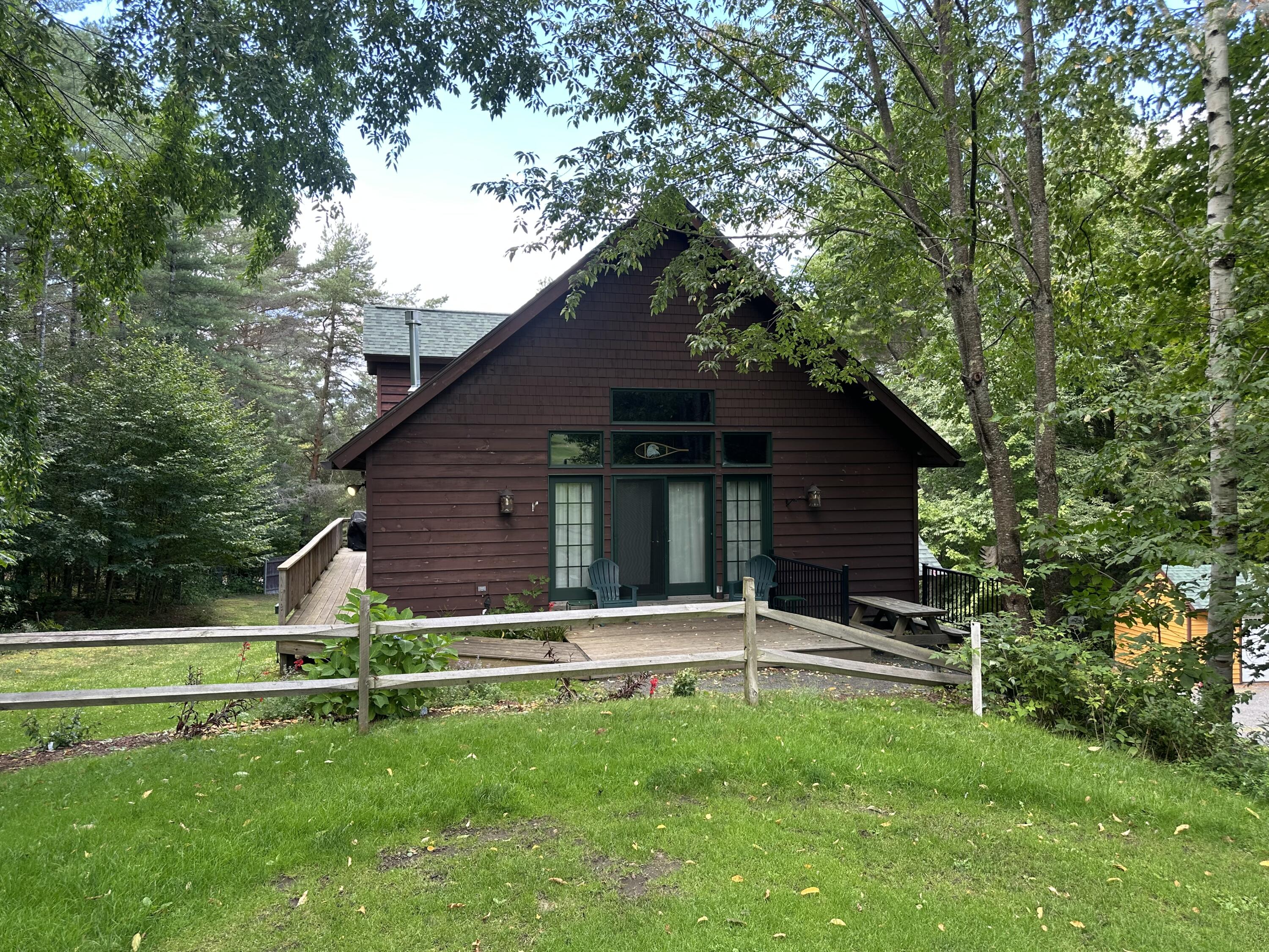 Dark brown gabled cabin nestled among trees, with a wooden deck and fenced lawn.