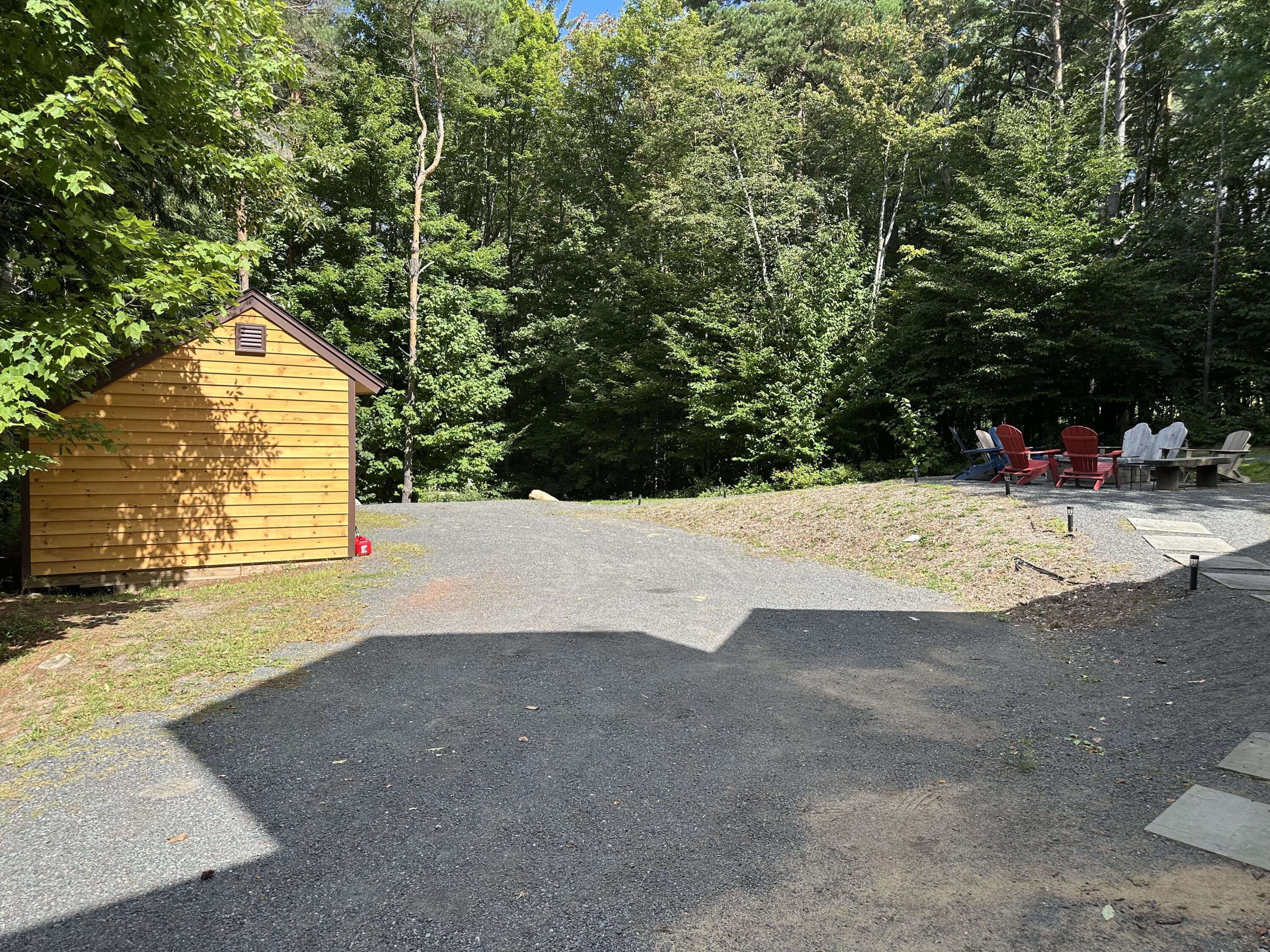 Gravel patio with a wooden shed and Adirondack chairs beside dense woods.