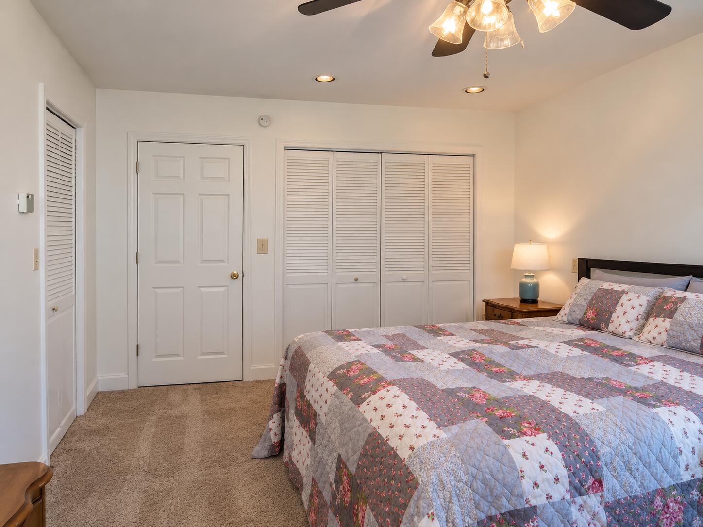 Bedroom with a floral patchwork quilt, louvered closet doors, and beige carpet.