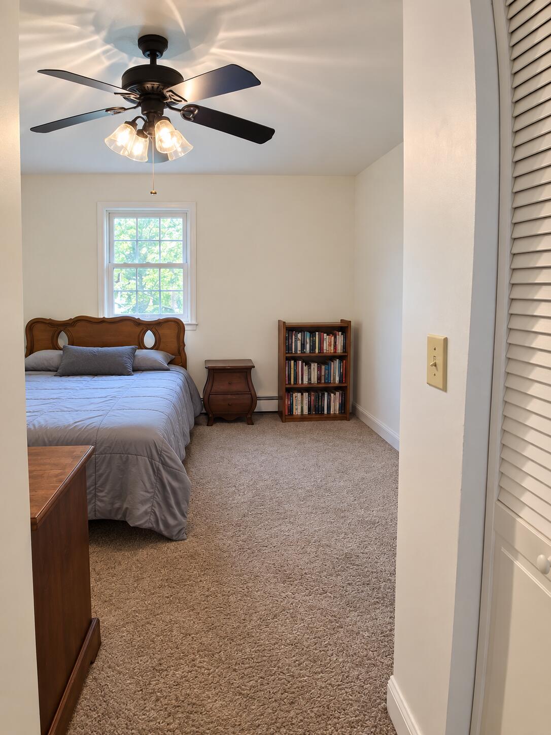 Furnished bedroom with gray bedding, a leafy view through the window, and a ceiling fan with lights.