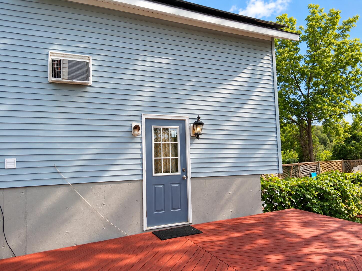 House exterior with a blue door opening onto a red wooden deck beside leafy trees.