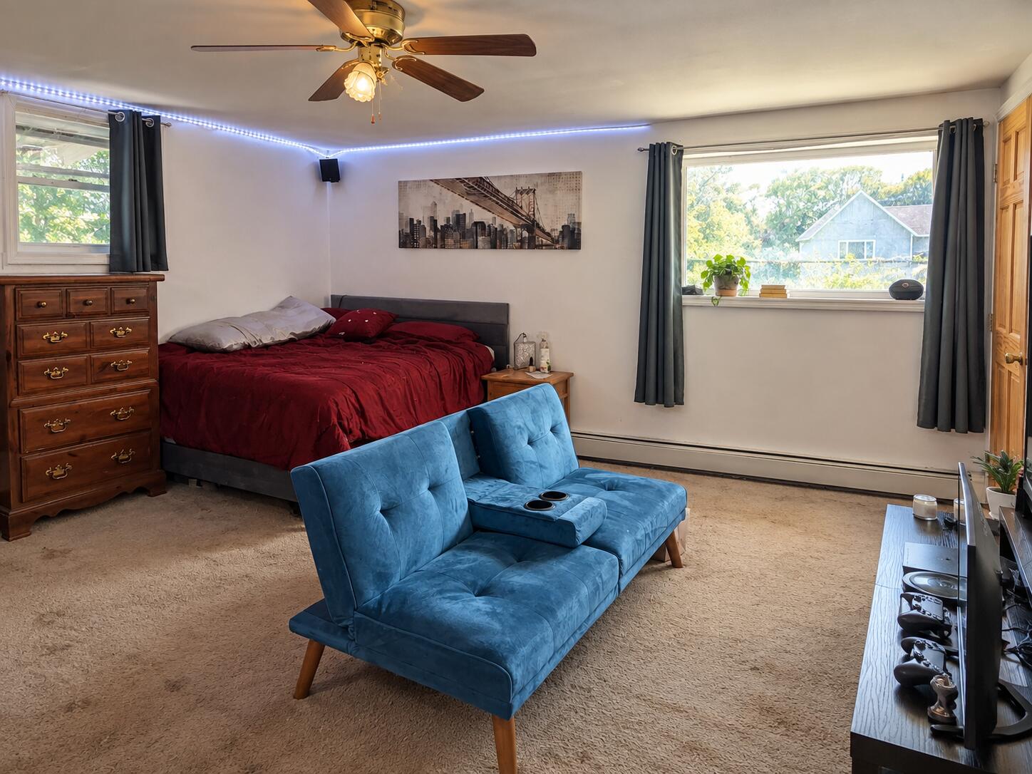 Bedroom with burgundy bedding, a blue loveseat, and windows overlooking neighboring homes.