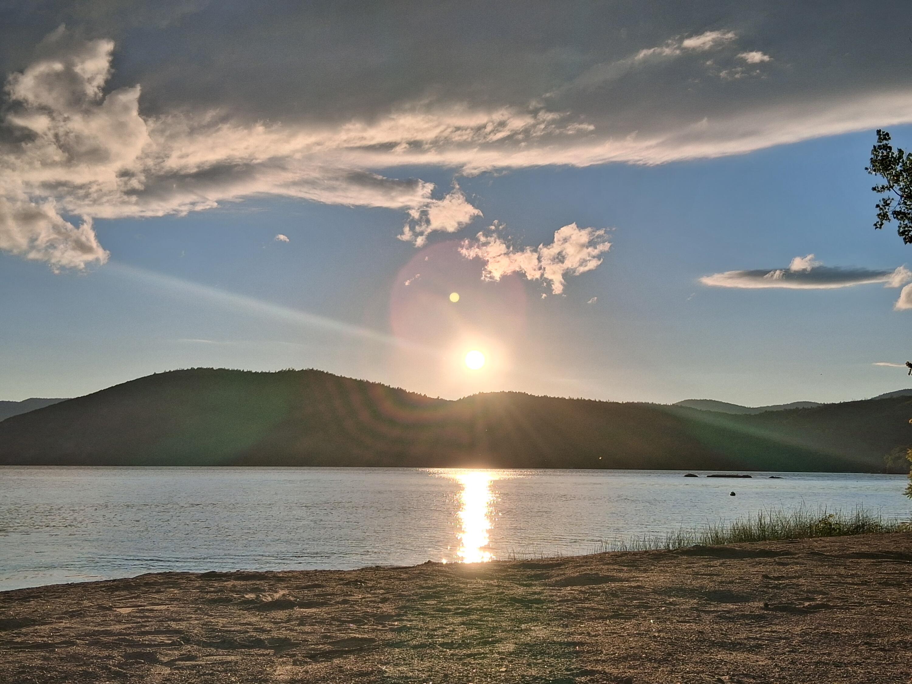 Sunset over a lake, with a bright reflection across the water and wooded hills beyond.