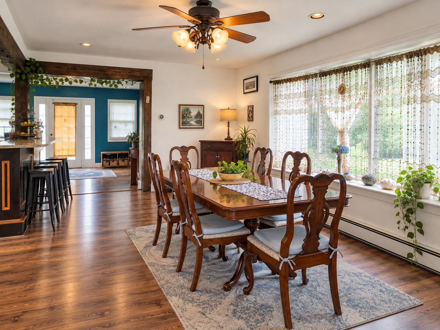 Dining room with a carved wood table beside a kitchen breakfast bar and a wall of curtained windows.