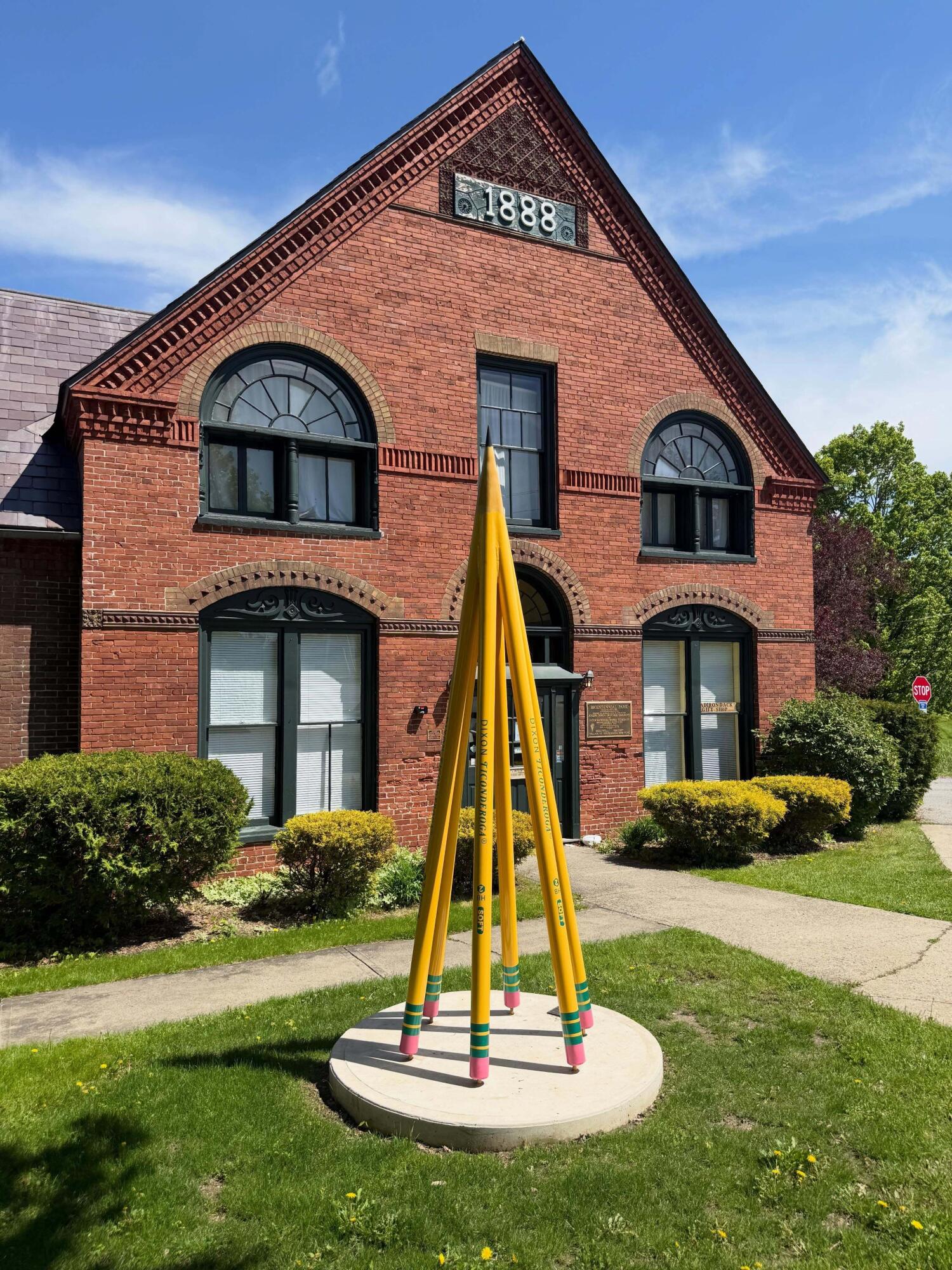Historic red-brick building with arched windows and a large yellow pencil sculpture on the lawn.