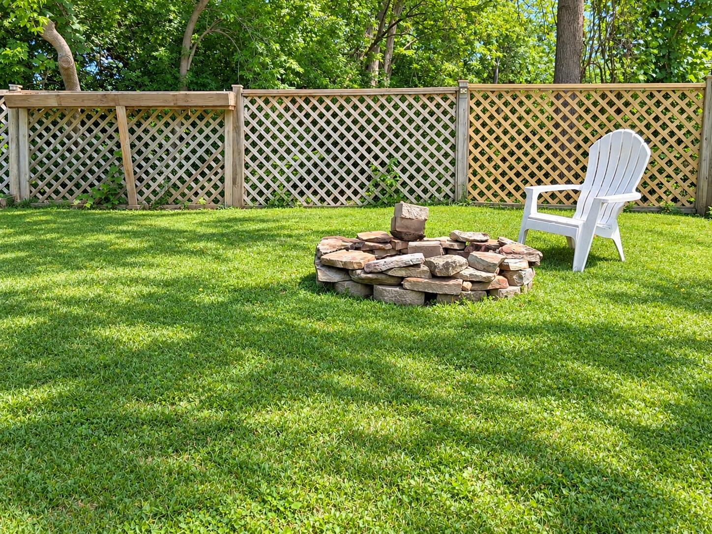 Backyard lawn with a stone fire pit and white Adirondack chair beside a lattice fence.